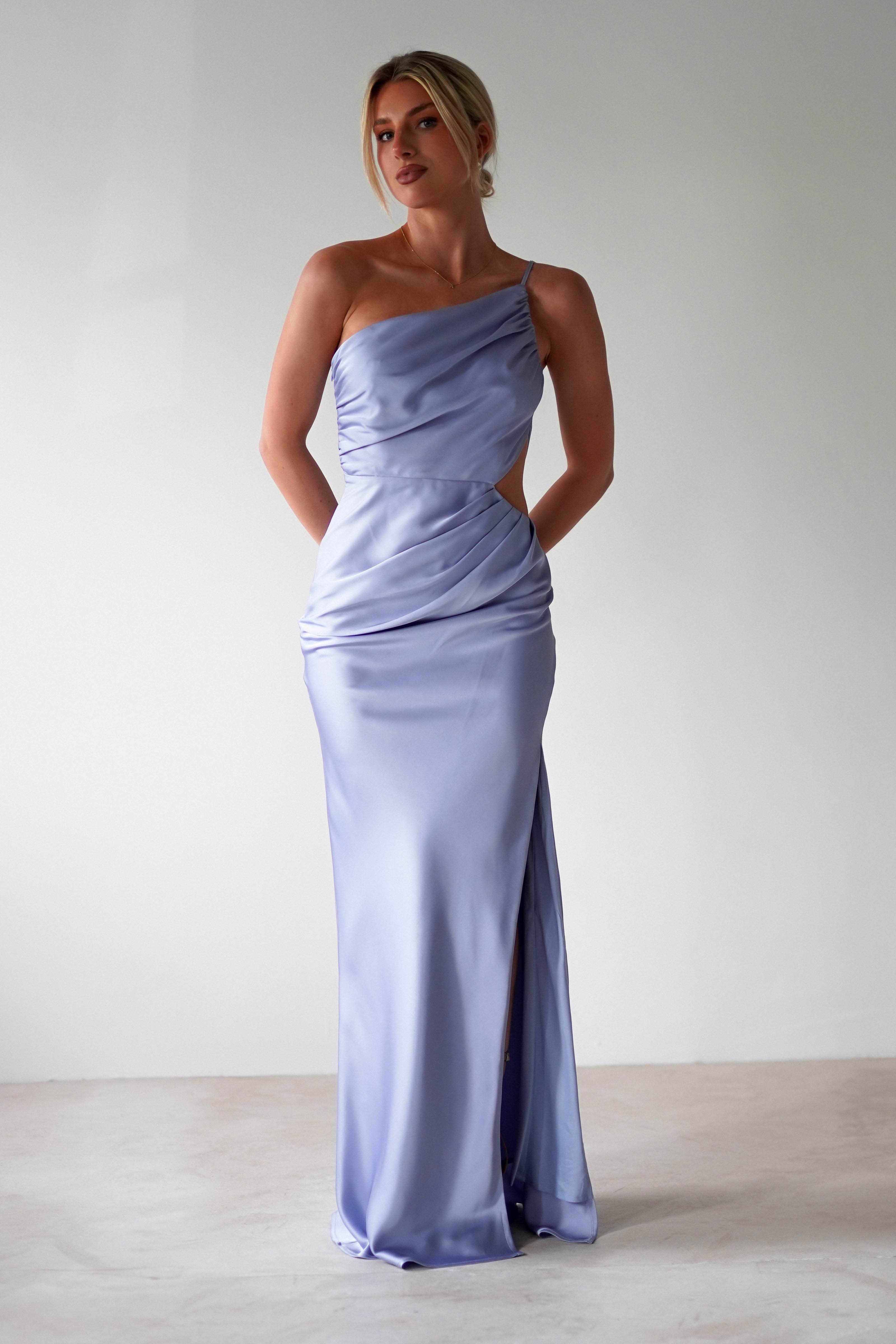 A woman stands against a plain light background, wearing a one-shoulder satin maxi gown in light purple with a fitted silhouette, hands behind her back—a perfect choice for bridesmaids.