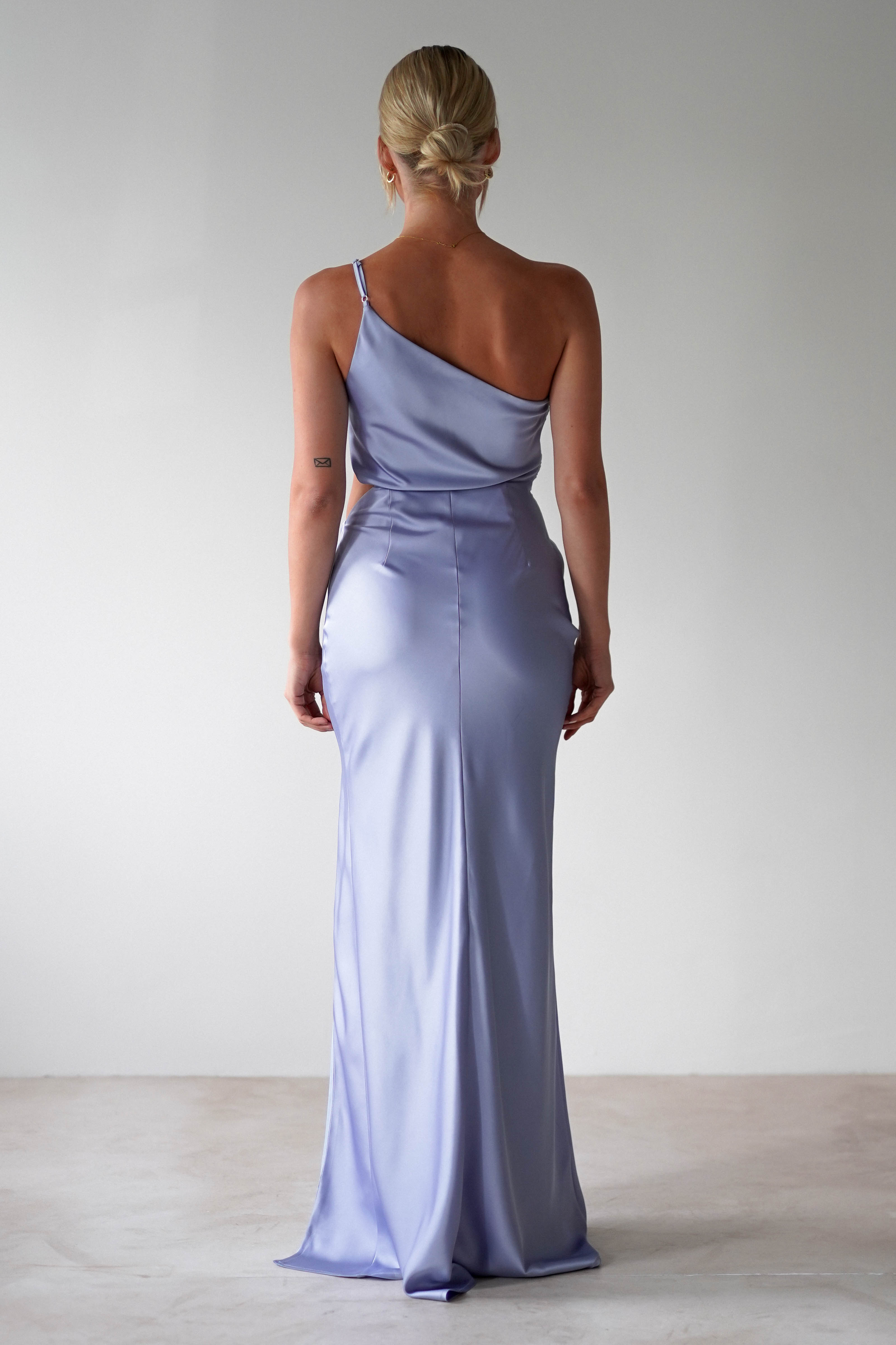 A woman with blonde hair in a low bun wears a shiny, light purple one-shoulder satin maxi gown, perfect for bridesmaids. She stands facing away from the camera against a plain, light background.
