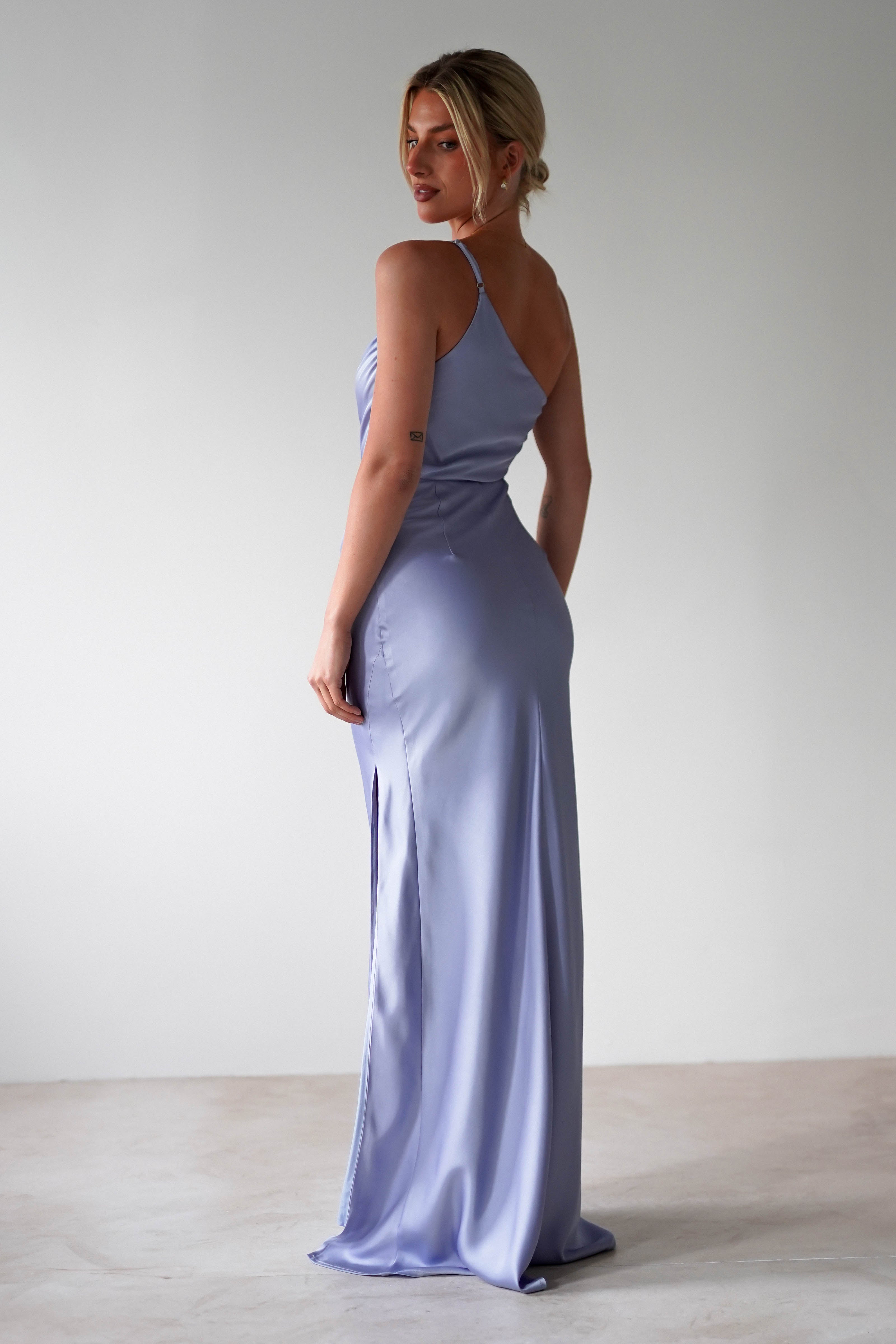 A woman stands in a minimalist room wearing an elegant one-shoulder satin maxi gown with a side slit—ideal for bridesmaids. She looks over her shoulder, her blonde hair styled in a low bun.