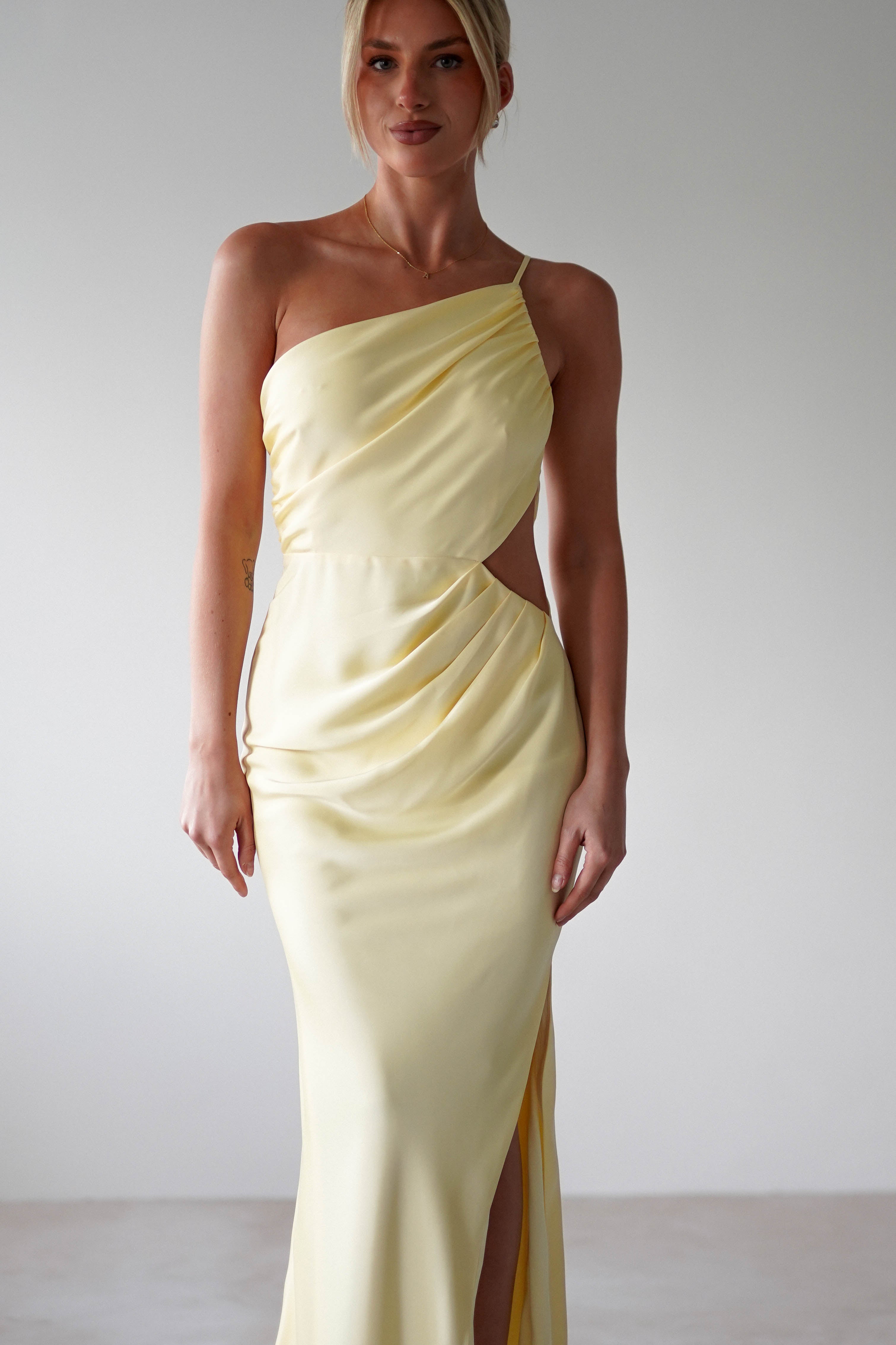 A woman wears a matte satin yellow one-shoulder maxi gown with a waist cutout and thigh-high slit, perfect for bridesmaids, standing against a plain, light background.