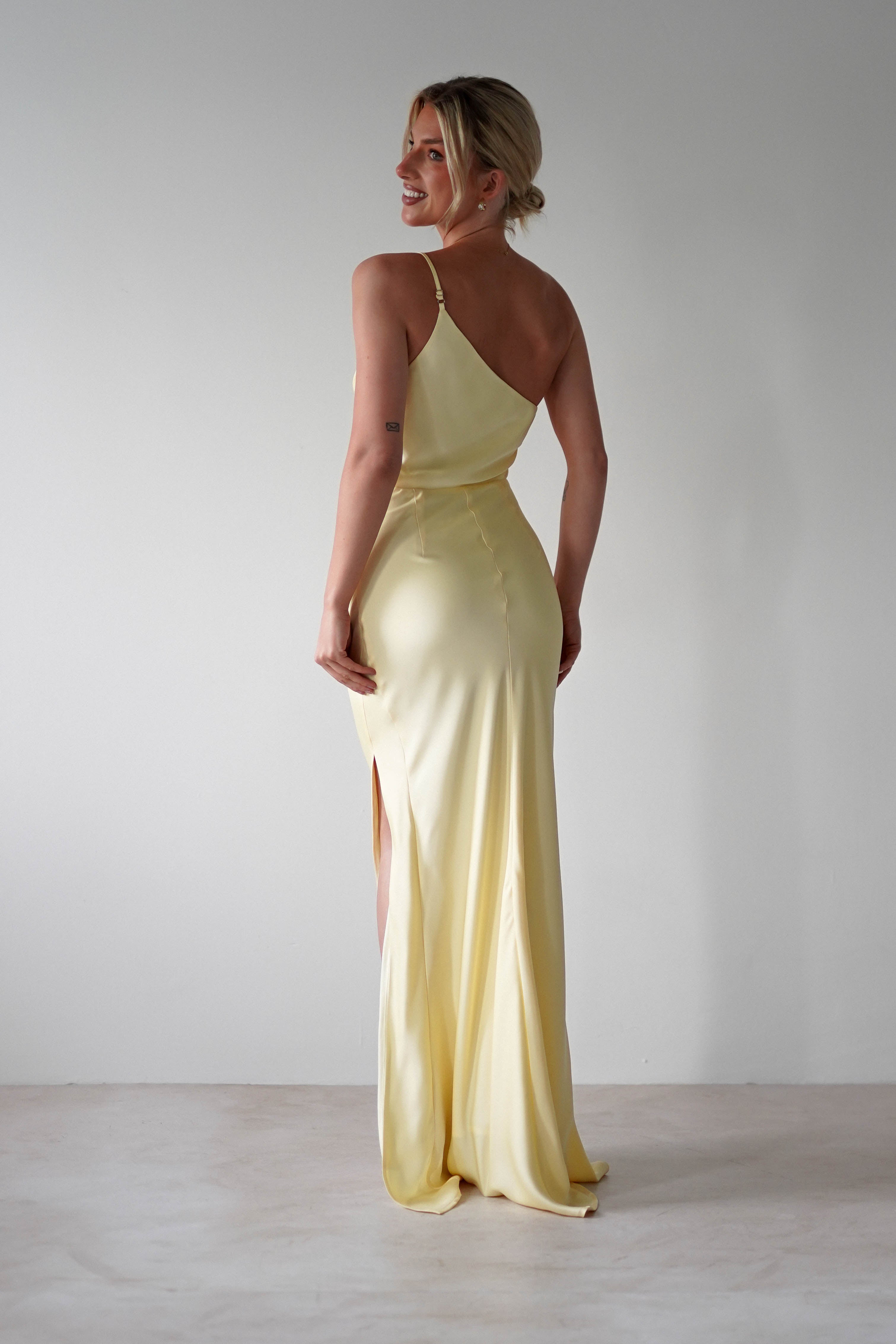 A woman in a matte satin yellow one-shoulder satin maxi dress, perfect for bridesmaids.