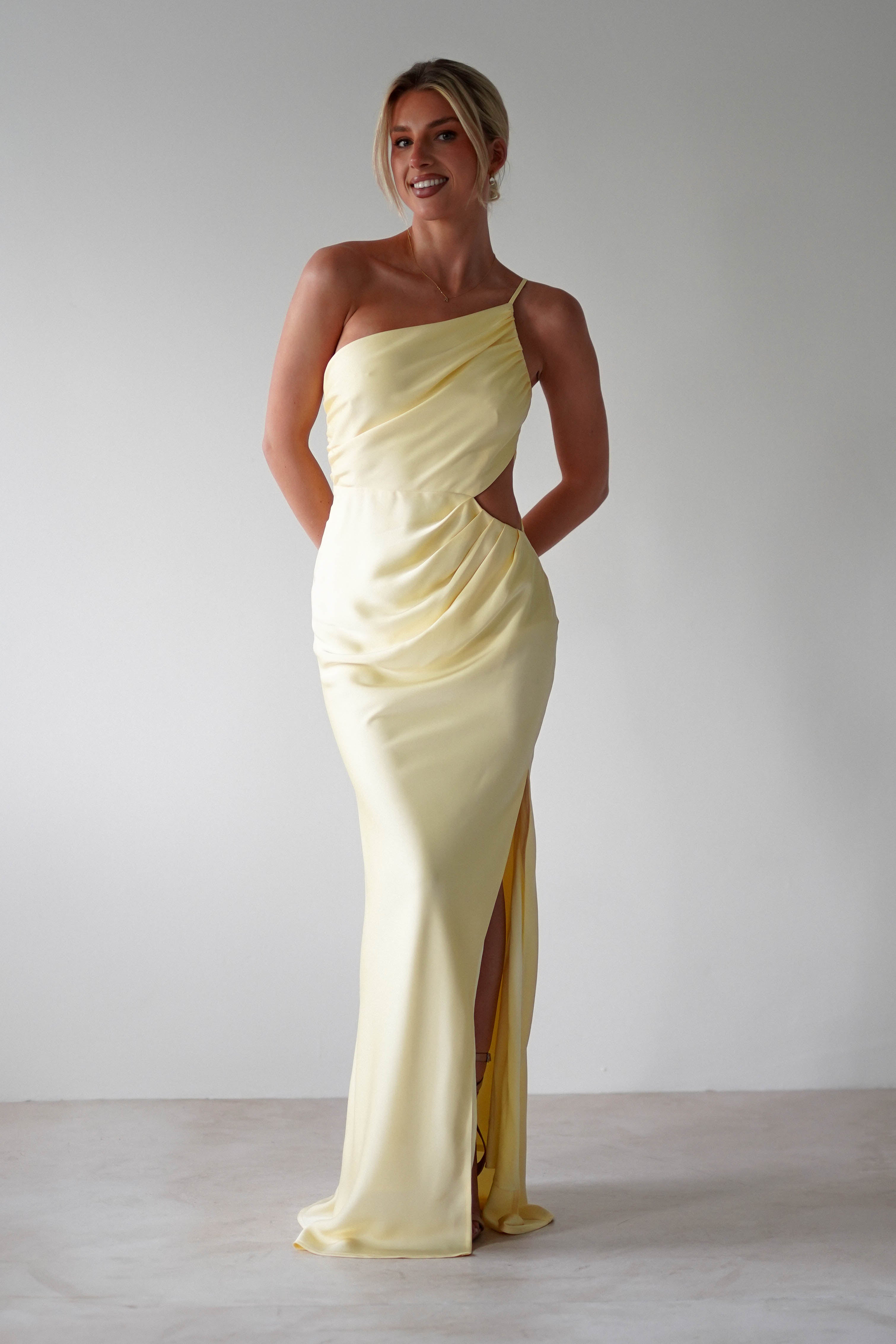 A woman smiles while wearing a pale yellow, one-shoulder satin maxi gown with side cutouts—an elegant choice for bridesmaids. She stands against a plain white background with her hands behind her back.