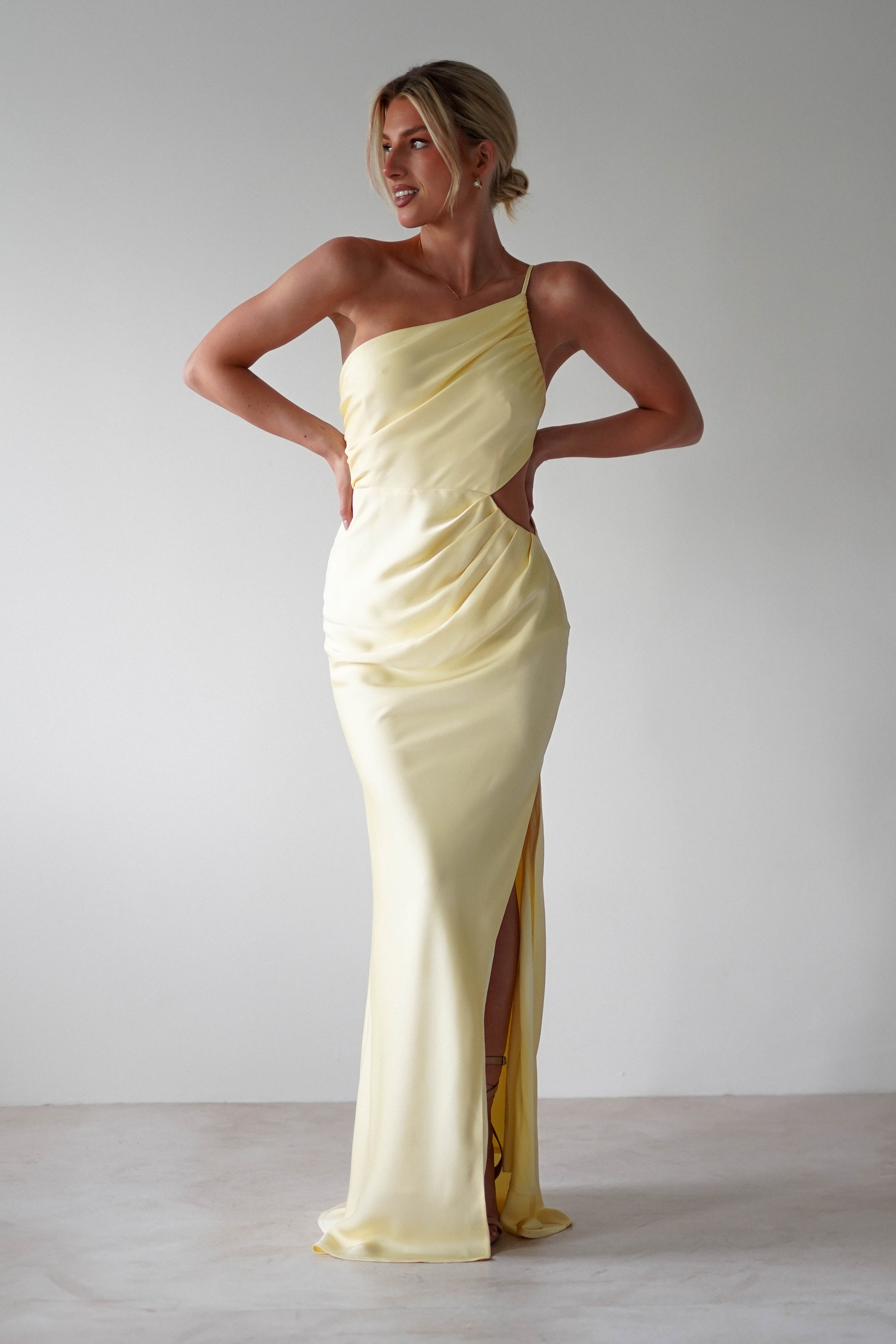 Woman wearing a matte satin yellow, one-shoulder satin maxi gown with a thigh-high slit, standing against a plain light background, hands on hips, looking to the side and smiling—perfect for bridesmaids.