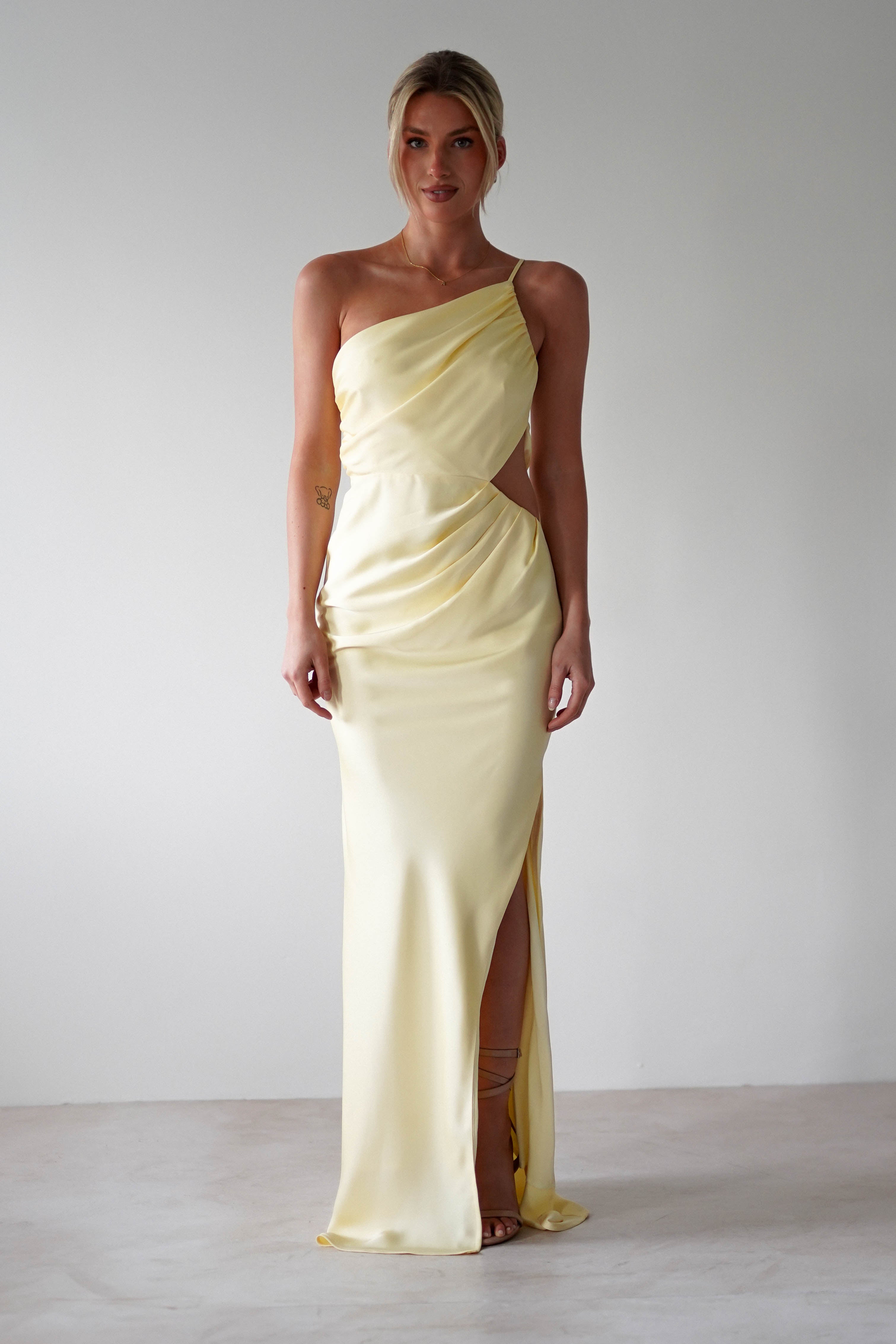 A woman stands against a plain background wearing a matte satin yellow, one-shoulder satin maxi gown with draped fabric and a thigh-high slit—an elegant choice for bridesmaids. Her blonde hair is styled in an updo as she faces forward.
