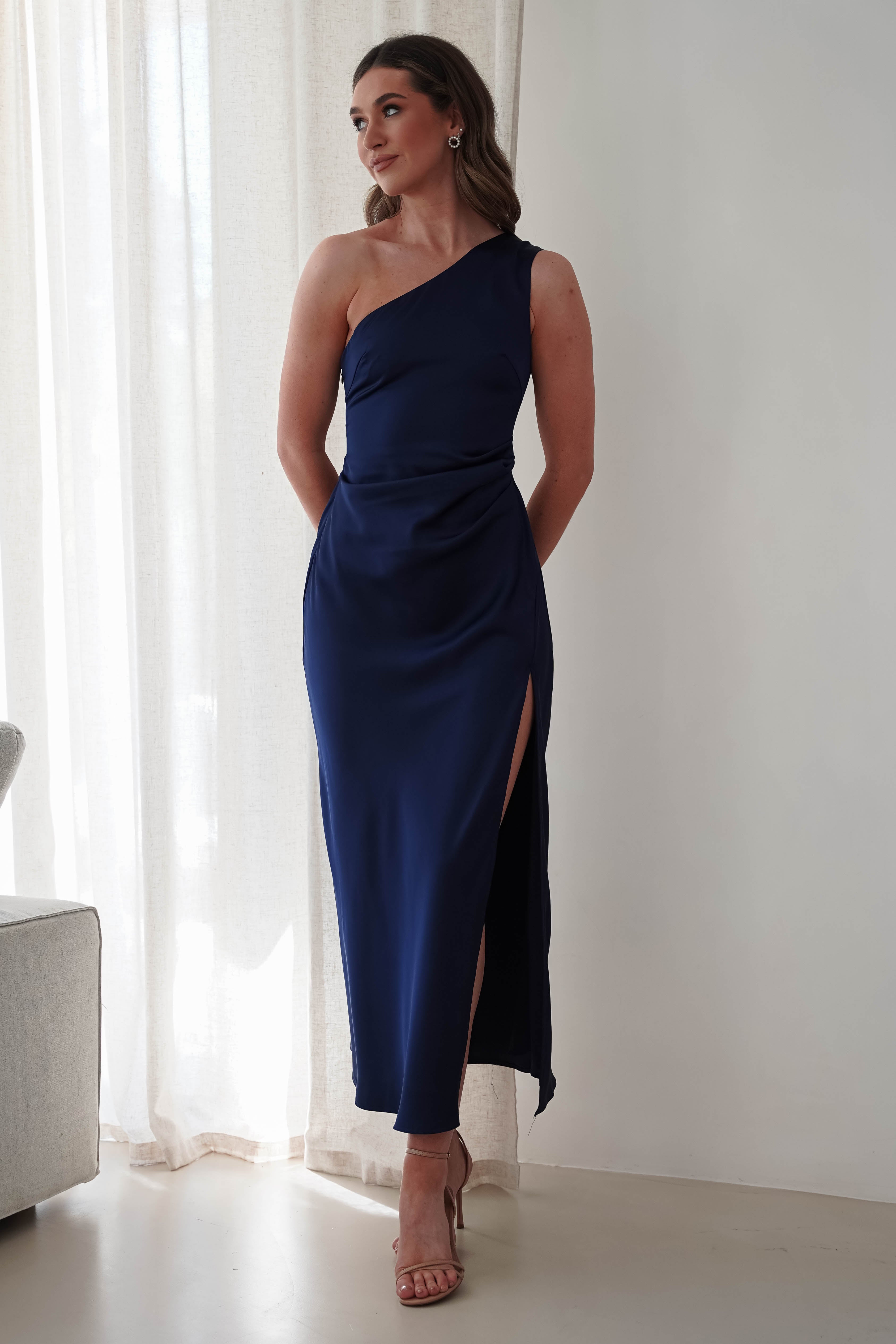 A person stands before a light curtain, wearing a navy blue wedding guest dress with a one-shoulder design and thigh-high slit, complemented by beige high heels. Their arms rest casually at their sides, while a light-colored sofa is partially visible on the left.