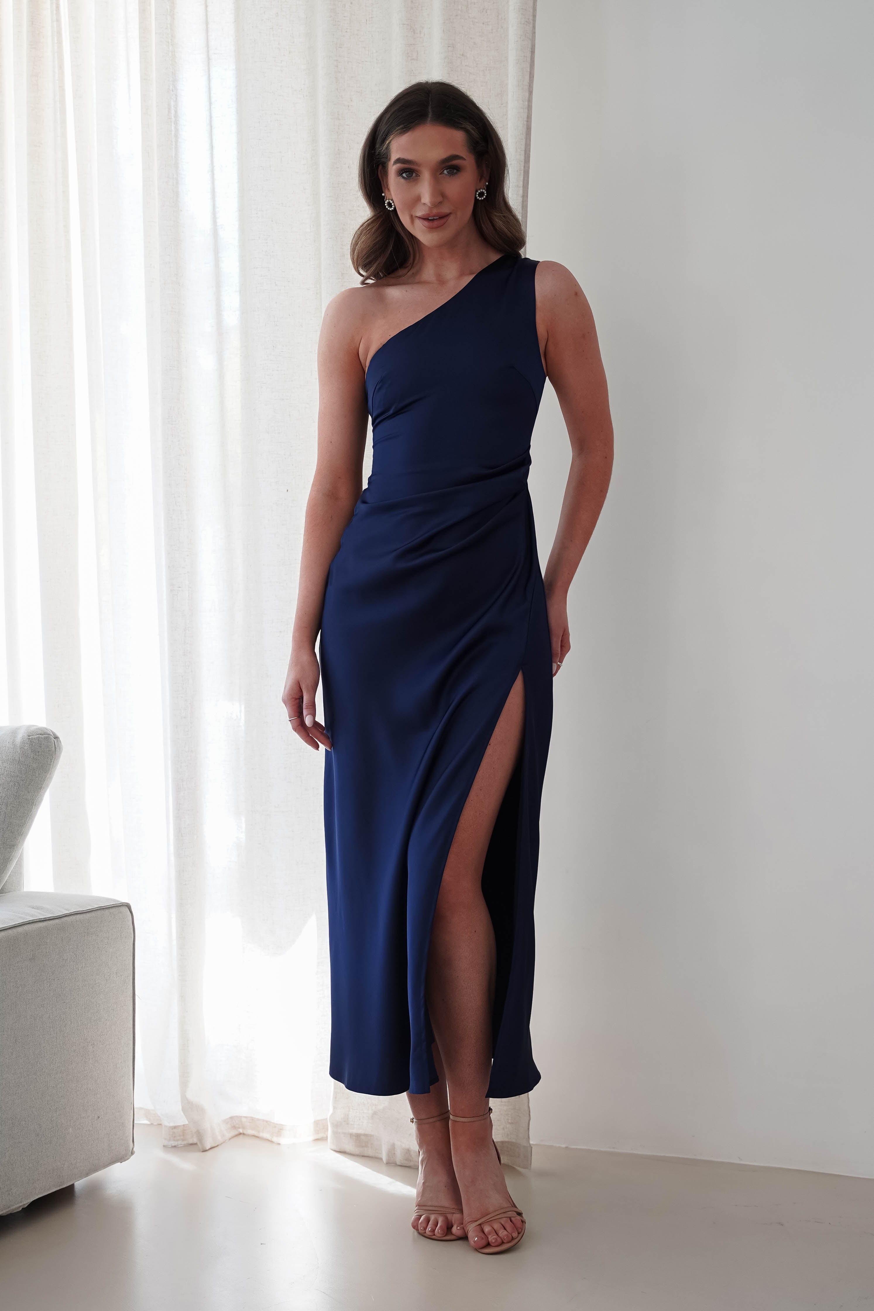 A woman in a one-shoulder, dark blue satin maxi dress with a thigh-high slit stands near a white curtain. She has long brown hair and is wearing strappy high heels. The room features a light, neutral color palette with part of a couch visible.