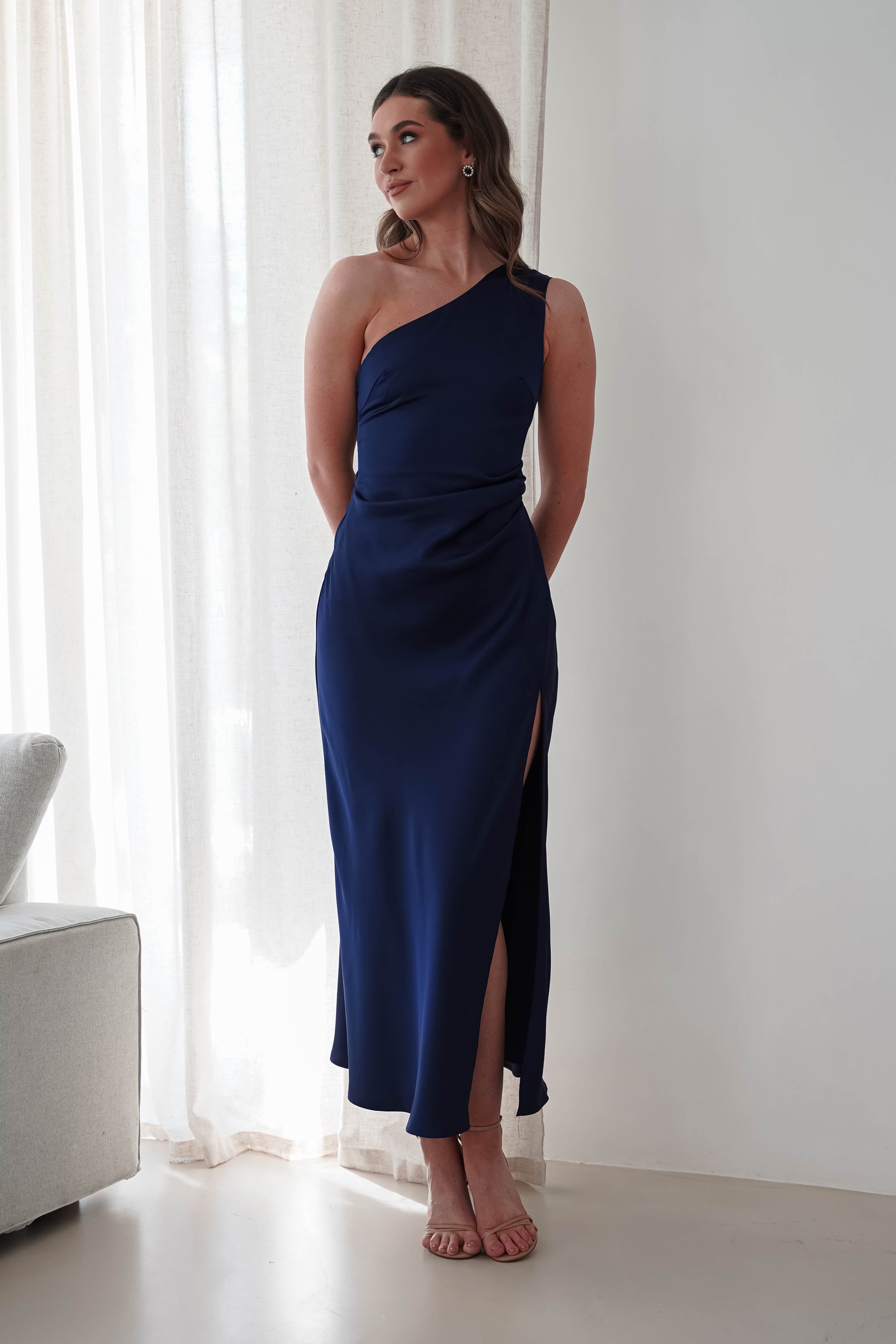 Woman in a blue satin maxi dress poses by a window with white curtains. The gown, perfect for a wedding guest or bridesmaid, features a thigh-high slit. She stands in a softly lit room with understated elegance, her hair styled simply and adorned with minimal accessories.