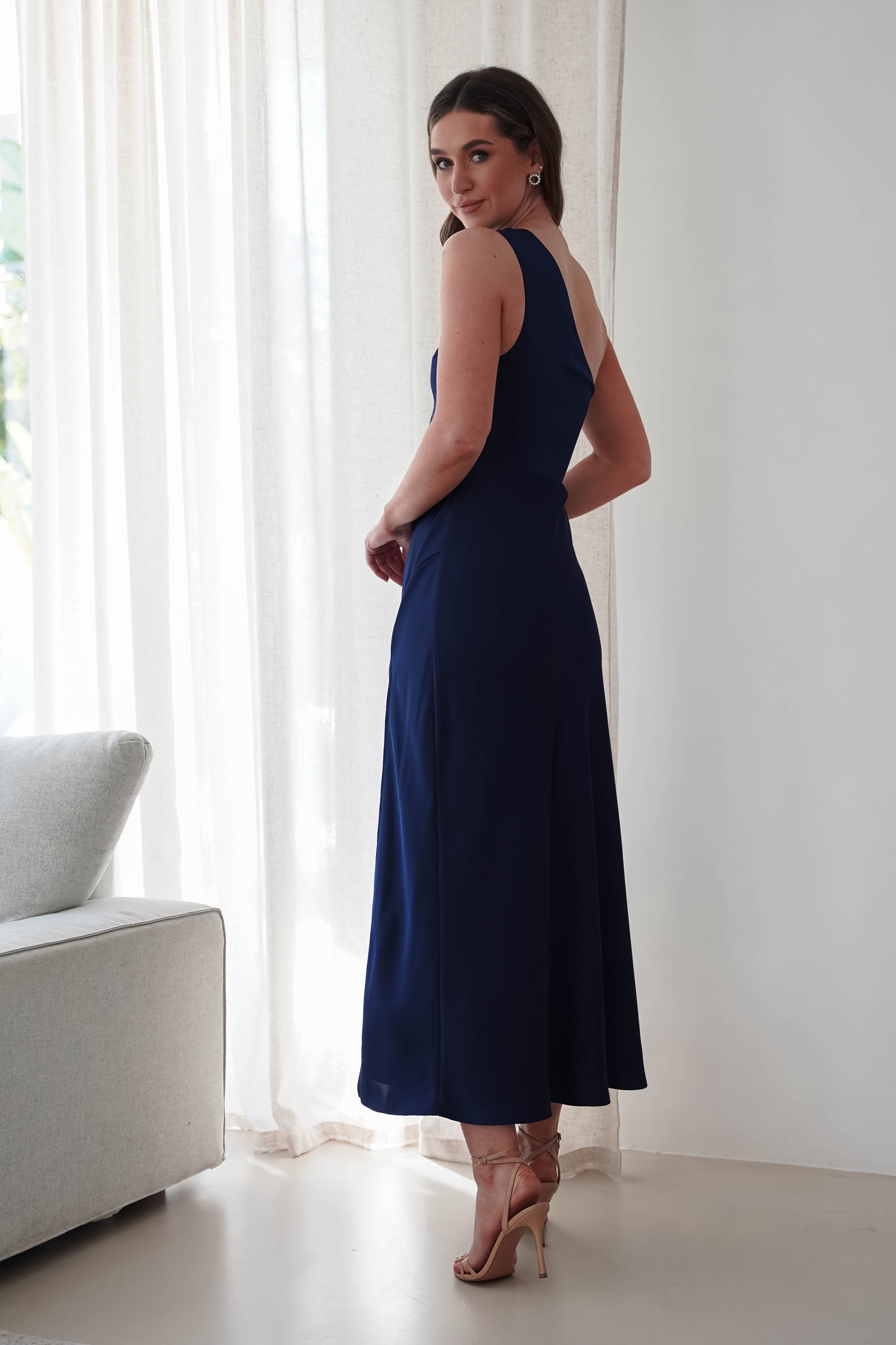 A woman in a sleeveless navy blue wedding guest dress stands beside a light gray sofa, facing a large window with sheer curtains. She is looking over her shoulder, with her hair pulled back, wearing high-heeled sandals.