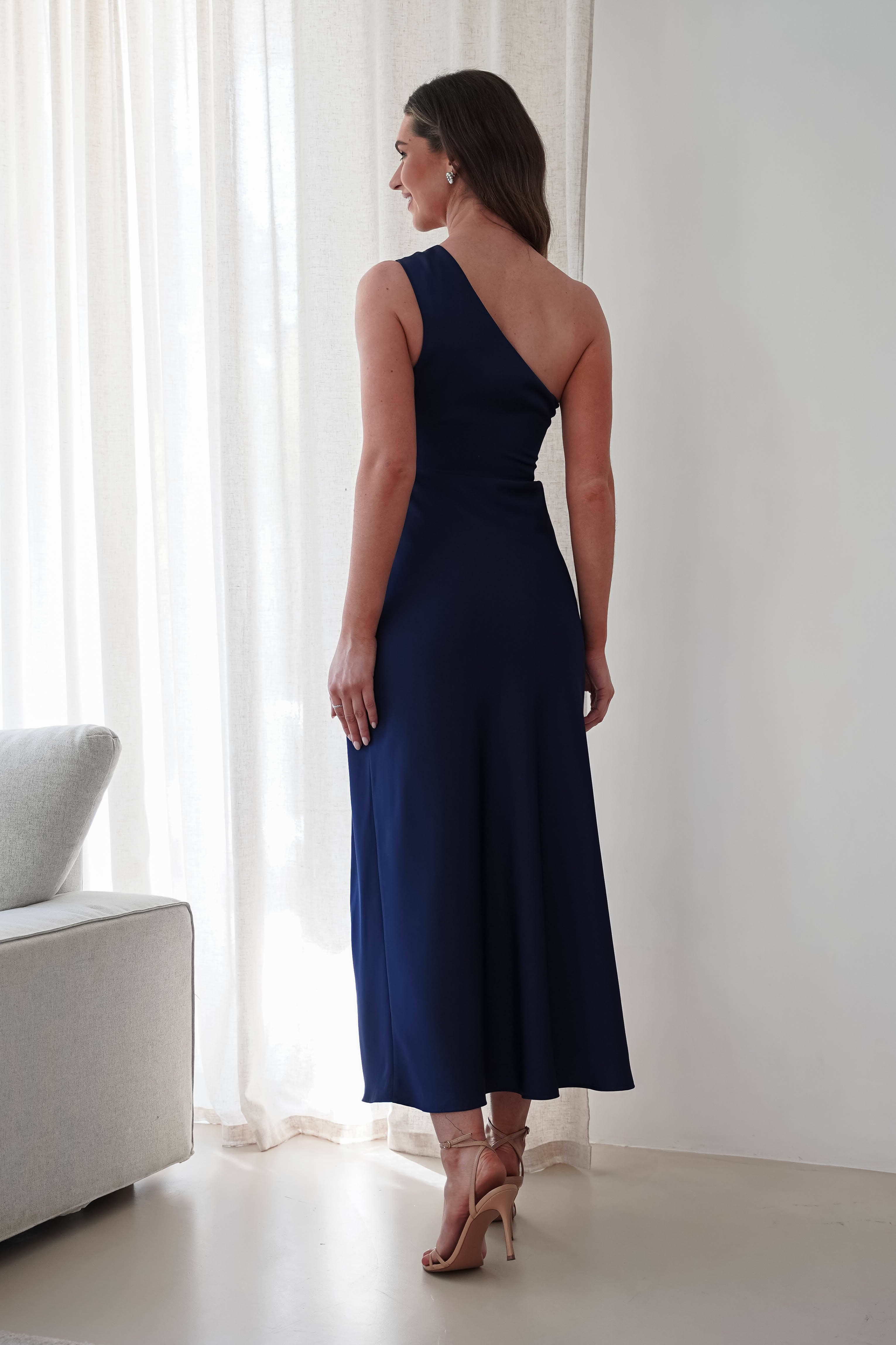 A woman stands in a room with her back to the camera, wearing a satin maxi dress and beige high heels. She is near a curtain beside a light-colored sofa, exuding elegance perfect for a wedding guest.