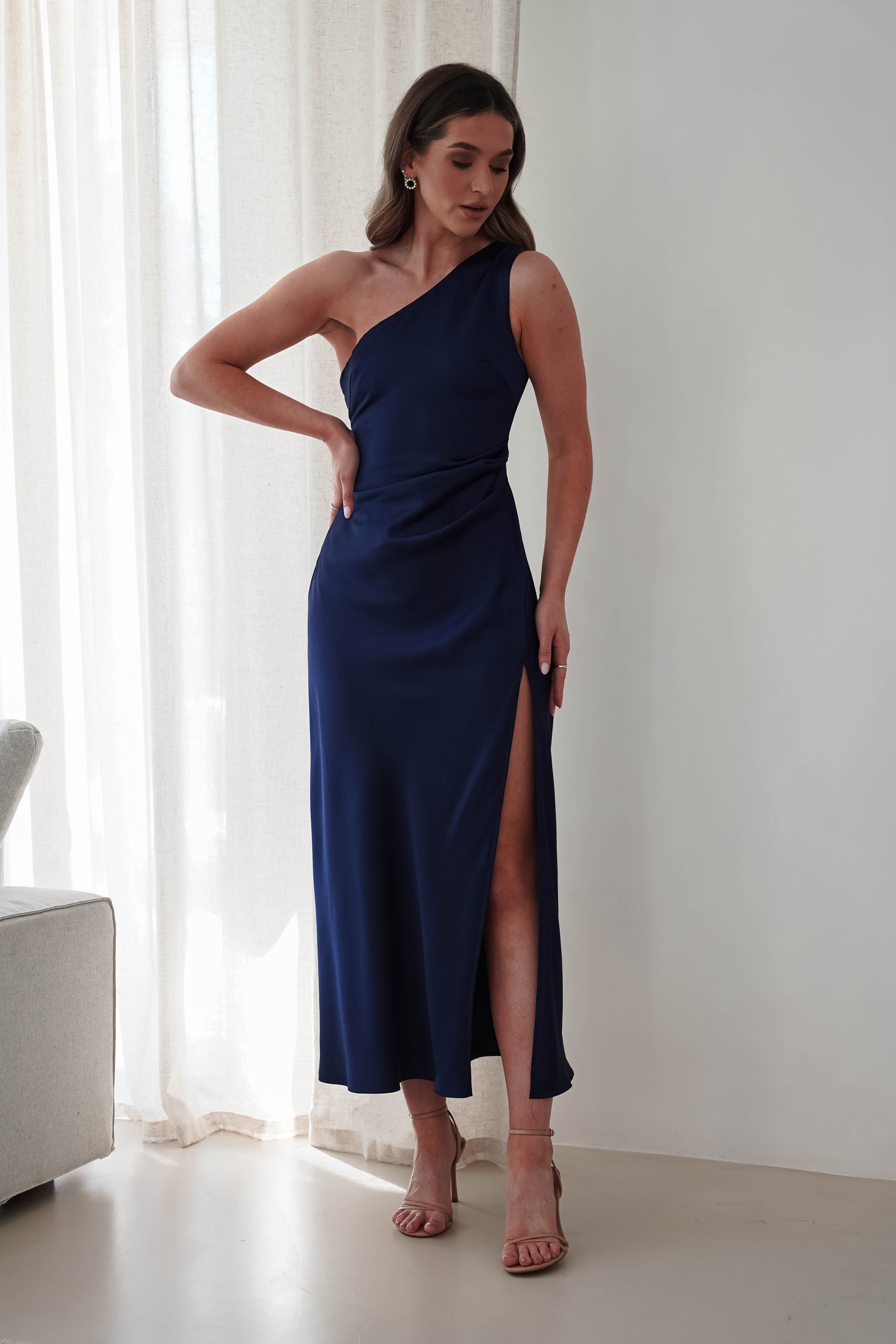 A woman stands in a room wearing a navy blue satin maxi dress with a side slit. She has long hair and is wearing heels, perfect as a wedding guest dress. The background features white curtains and a light-colored sofa.