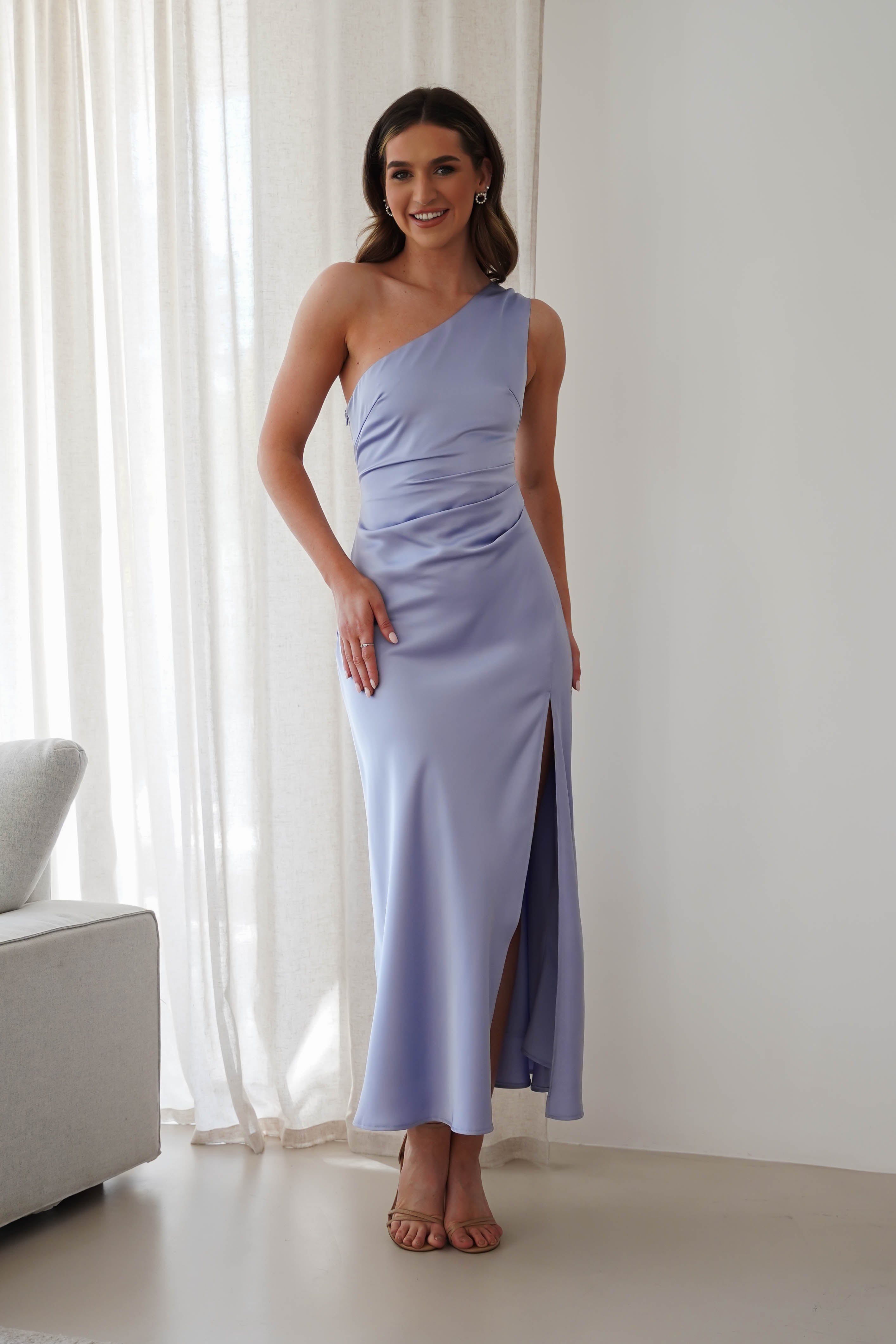 A woman stands in a light-filled room wearing a sleek, light purple bridesmaid dress with a thigh-high slit. She has long hair and is smiling. A white curtain and part of a gray couch are visible in the background.