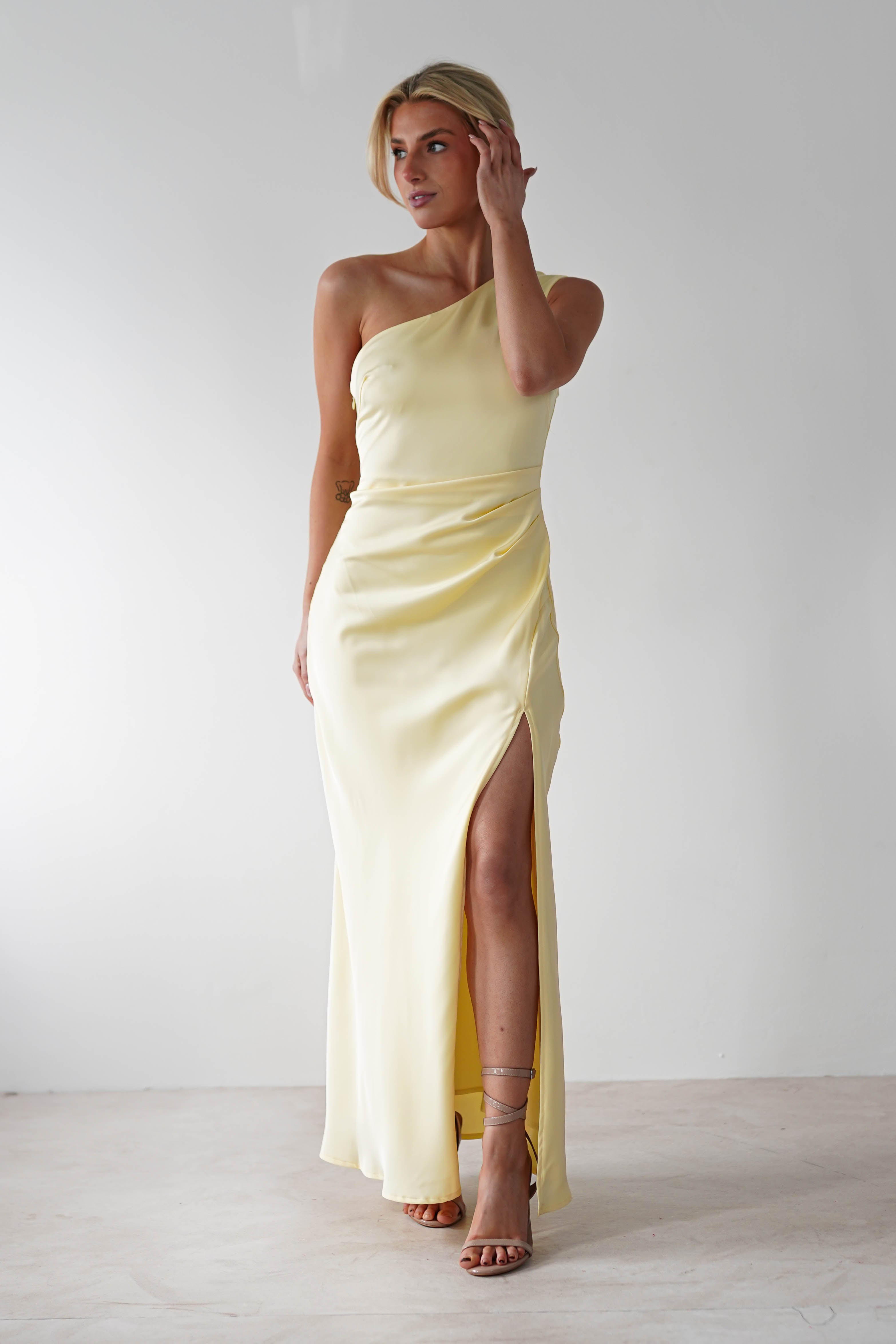 A person poses in a pale yellow, one-shoulder satin maxi dress with a thigh-high slit. They have short blonde hair and wear strappy sandals. The background is a plain, light-colored wall.