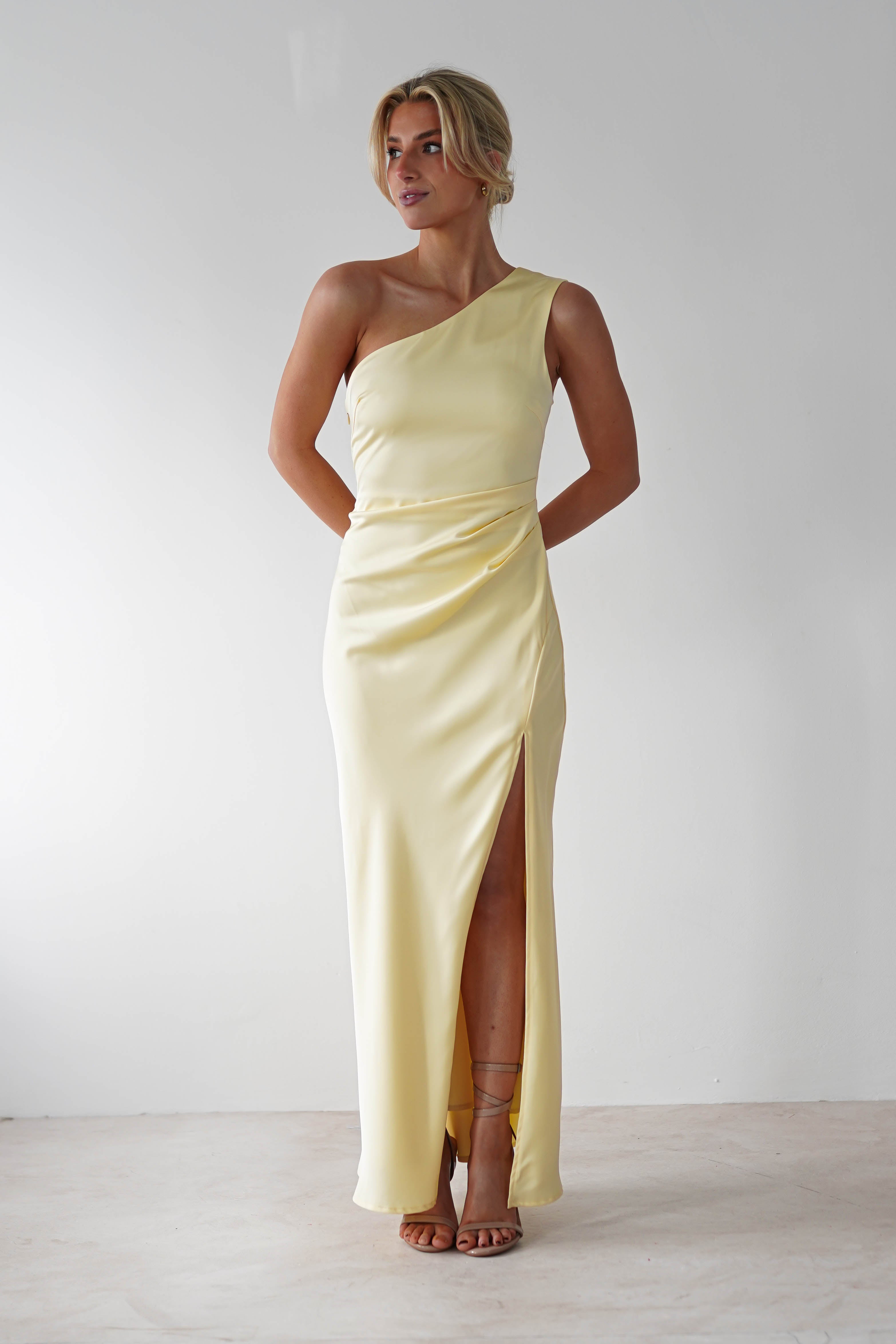 A person wearing a light yellow one-shoulder satin maxi dress with a thigh-high slit stands against a plain white background. The dress, perfect for a wedding guest, drapes elegantly with a soft sheen as the person poses with hands behind their back.