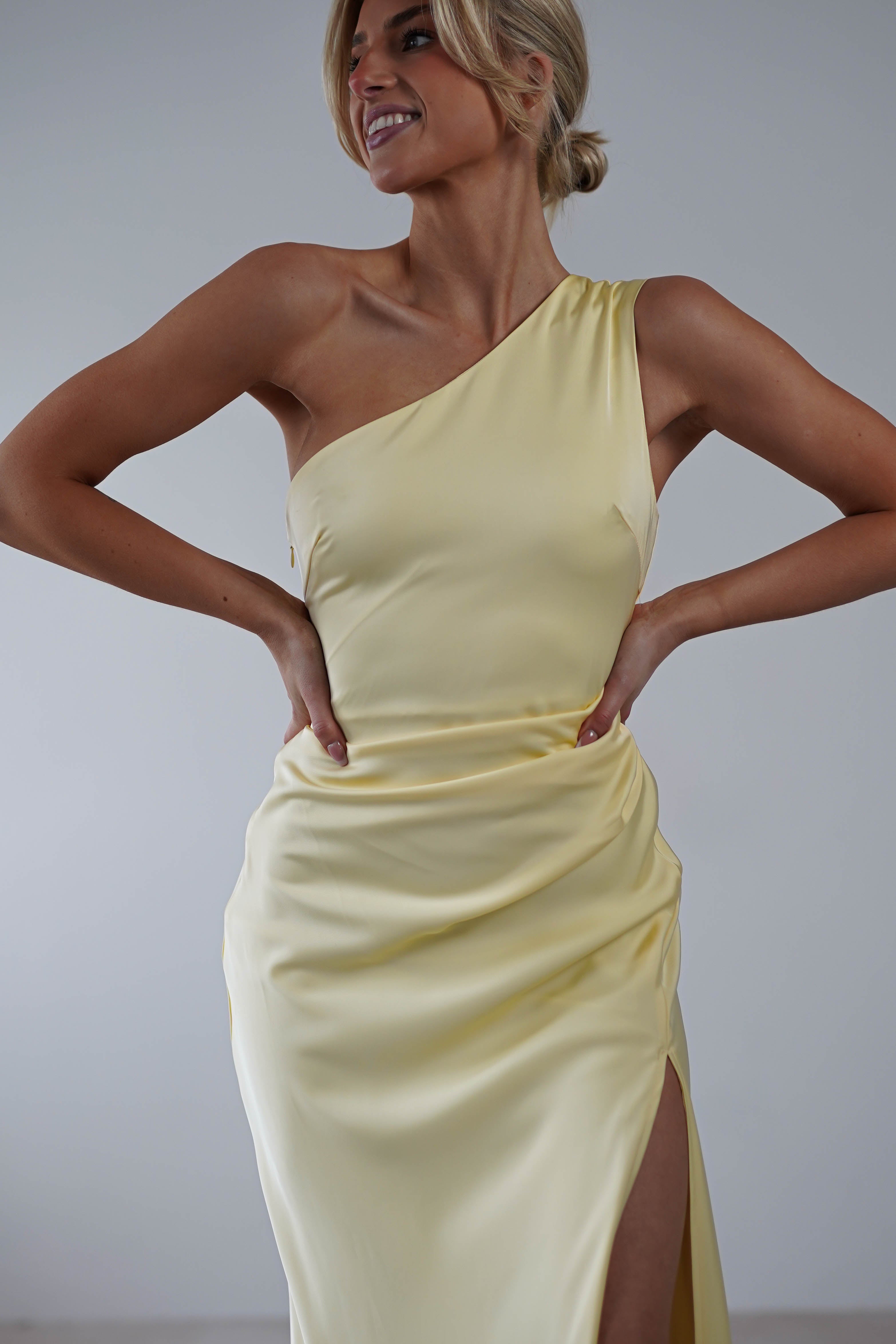 A woman in a pale yellow, one-shoulder bridesmaid dress stands with her hands on her hips. The dress has a slit on one side, and she has short, styled hair. The background is plain gray.