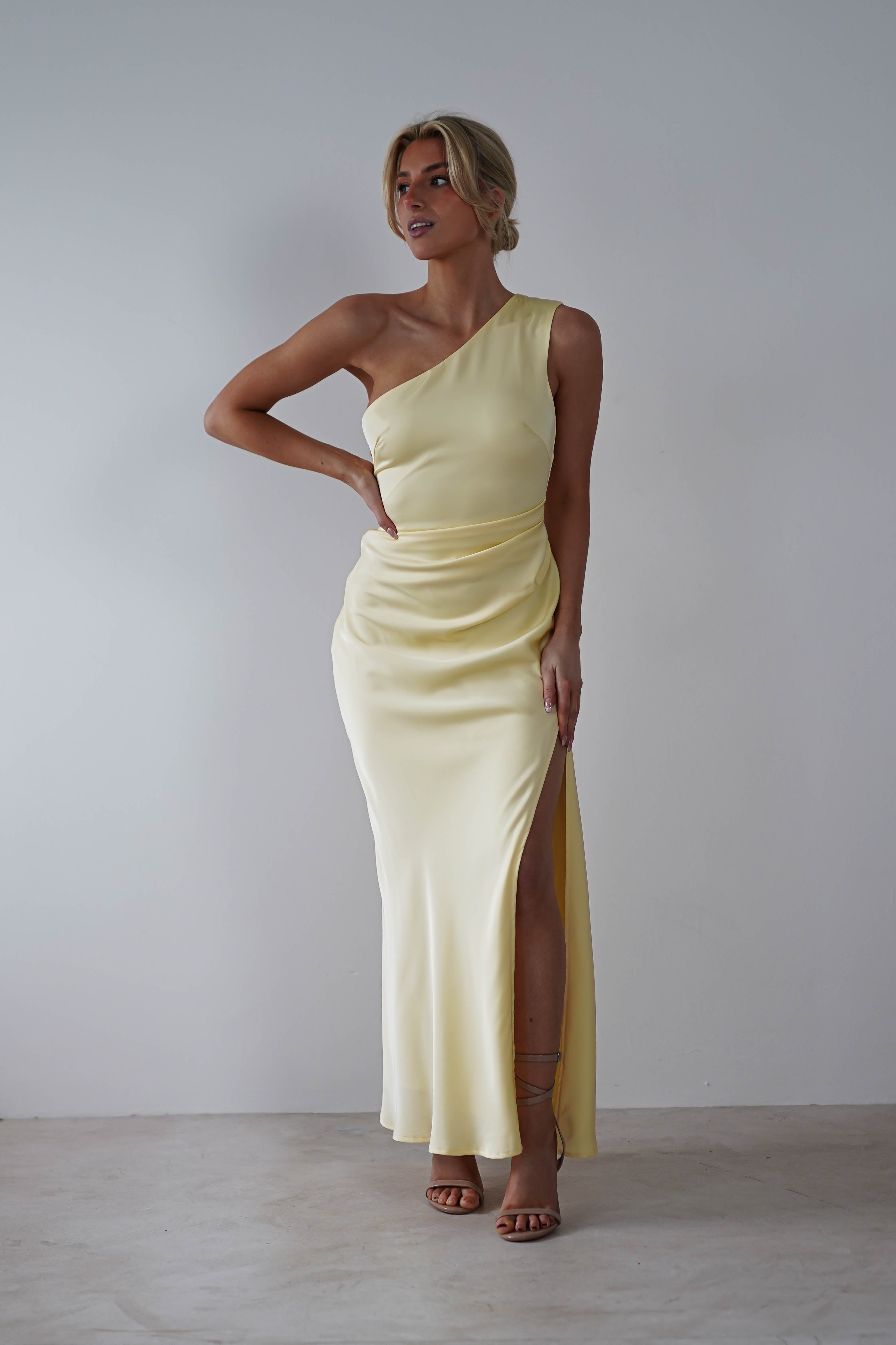 A person wearing a one-shoulder satin maxi dress with a high slit stands gracefully against a plain background, exuding elegance with one hand on their hip and hair styled in an updo—a perfect look for a wedding guest or bridesmaid.