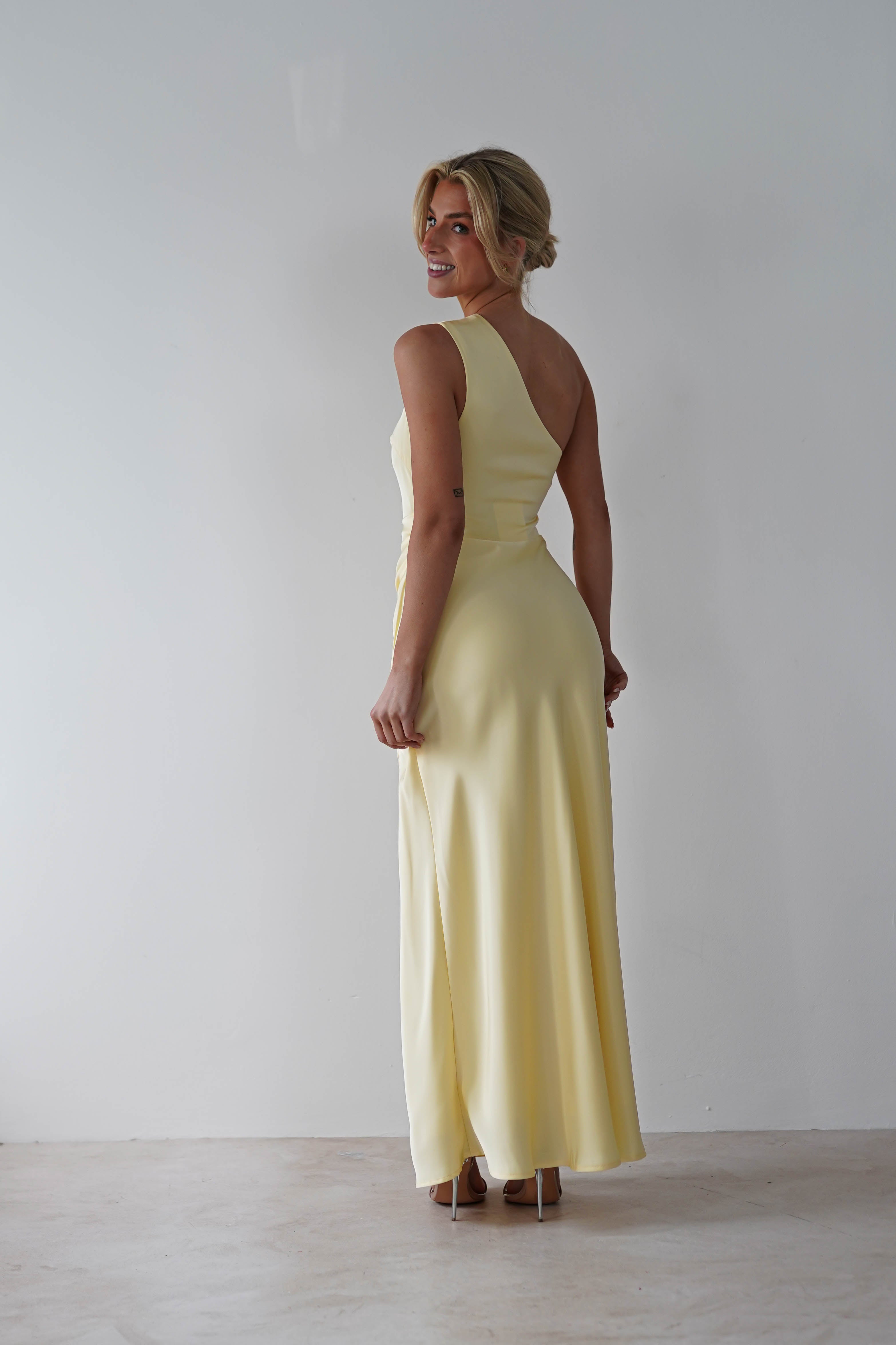 A woman in a long, light yellow wedding guest dress stands against a white background, looking over her shoulder and smiling. Her hair is styled in an updo, perfectly complementing her elegant look. She wears stylish two-tone platform shoes.