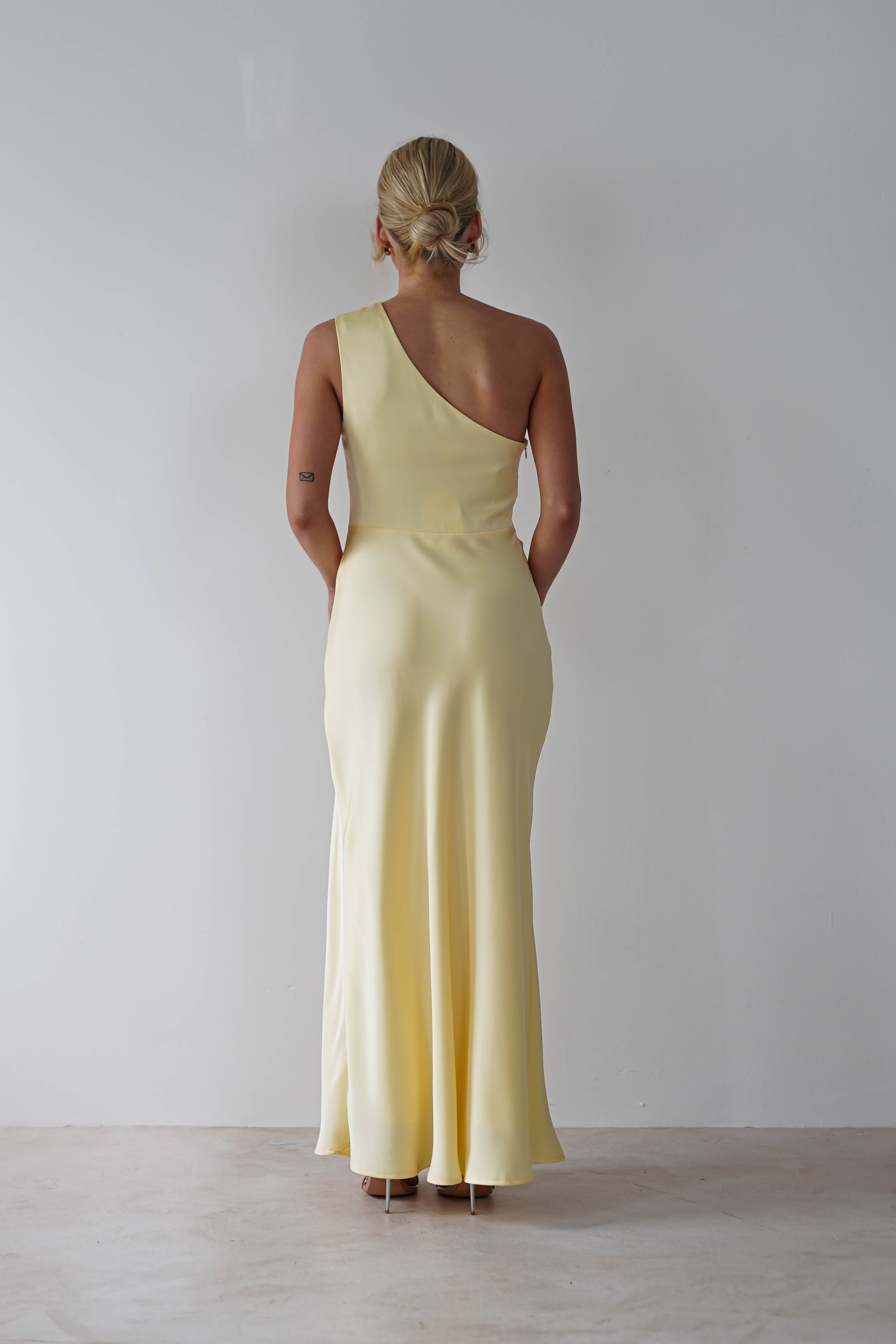 A person with blonde hair stands with their back to the camera, draped in a stunning one-shoulder satin maxi dress in light yellow, set against a plain white background.