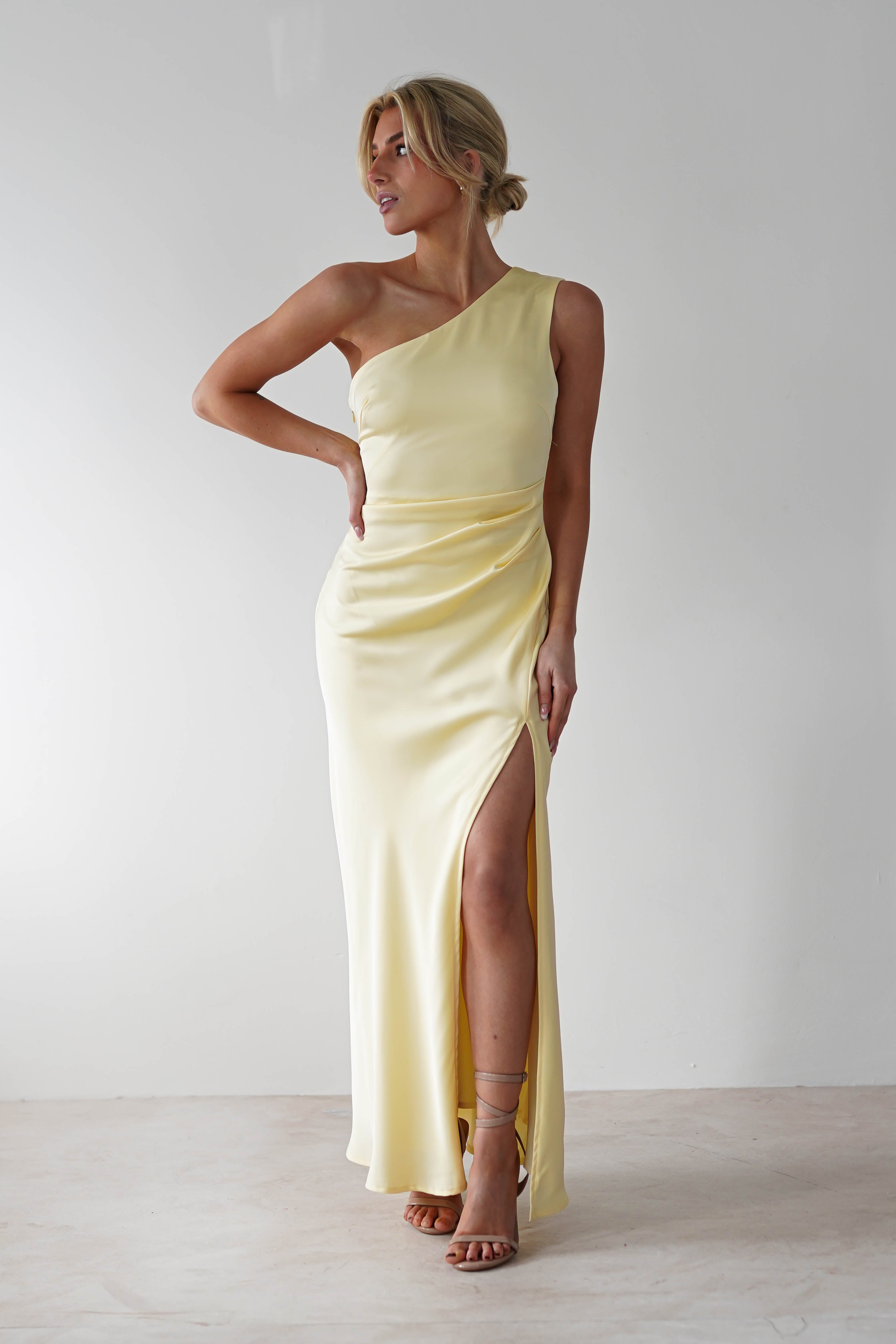 A person wearing a light yellow one-shoulder satin maxi dress with a thigh-high slit stands against a plain white background. They have one hand on their hip and the other resting by their side, gazing to the right with an elegant pose suitable for a wedding guest dress.