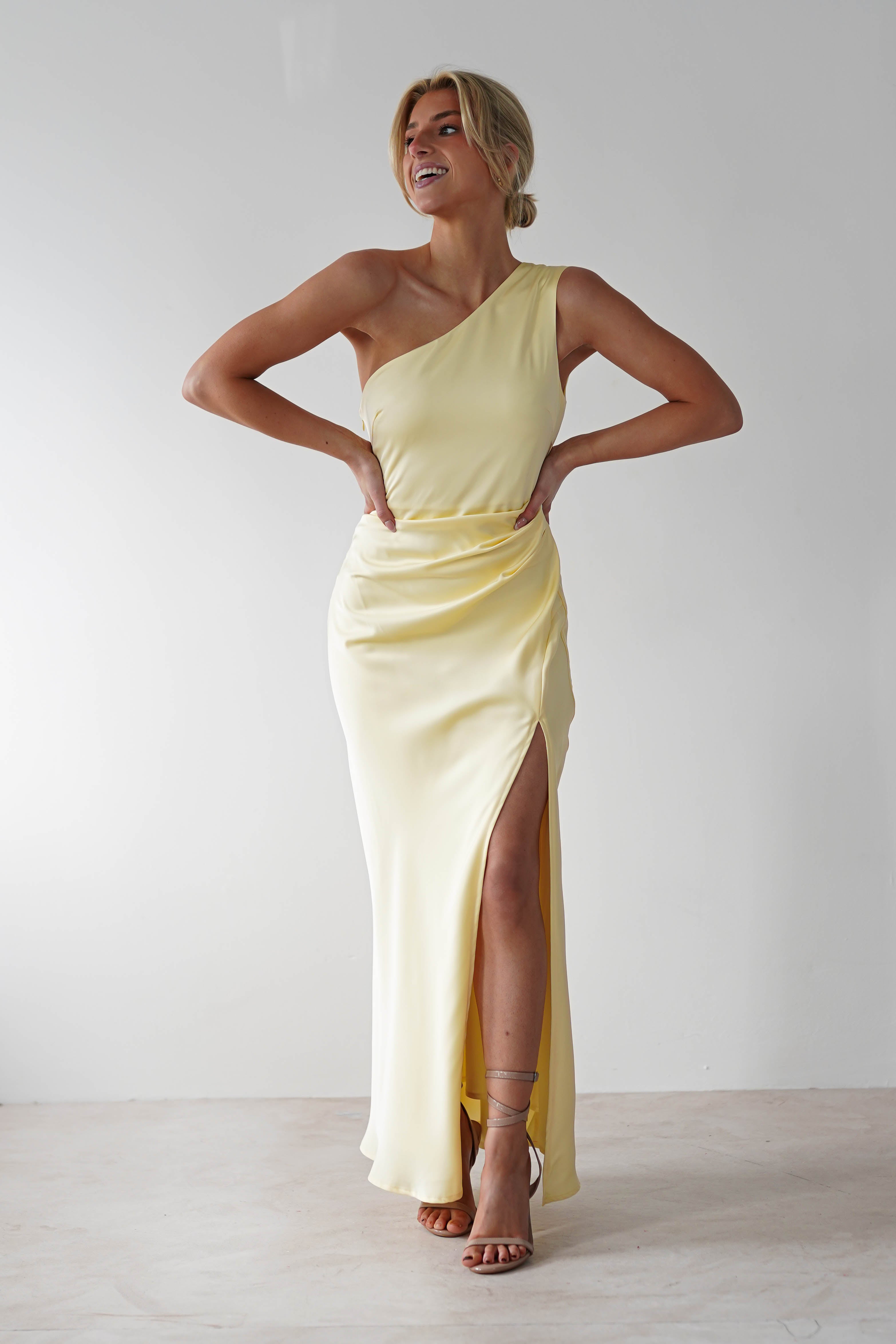 A person stands confidently against a plain background, wearing a sleeveless yellow bridesmaid dress with an asymmetrical neckline and a thigh-high slit. They have their hands on their hips and are smiling.