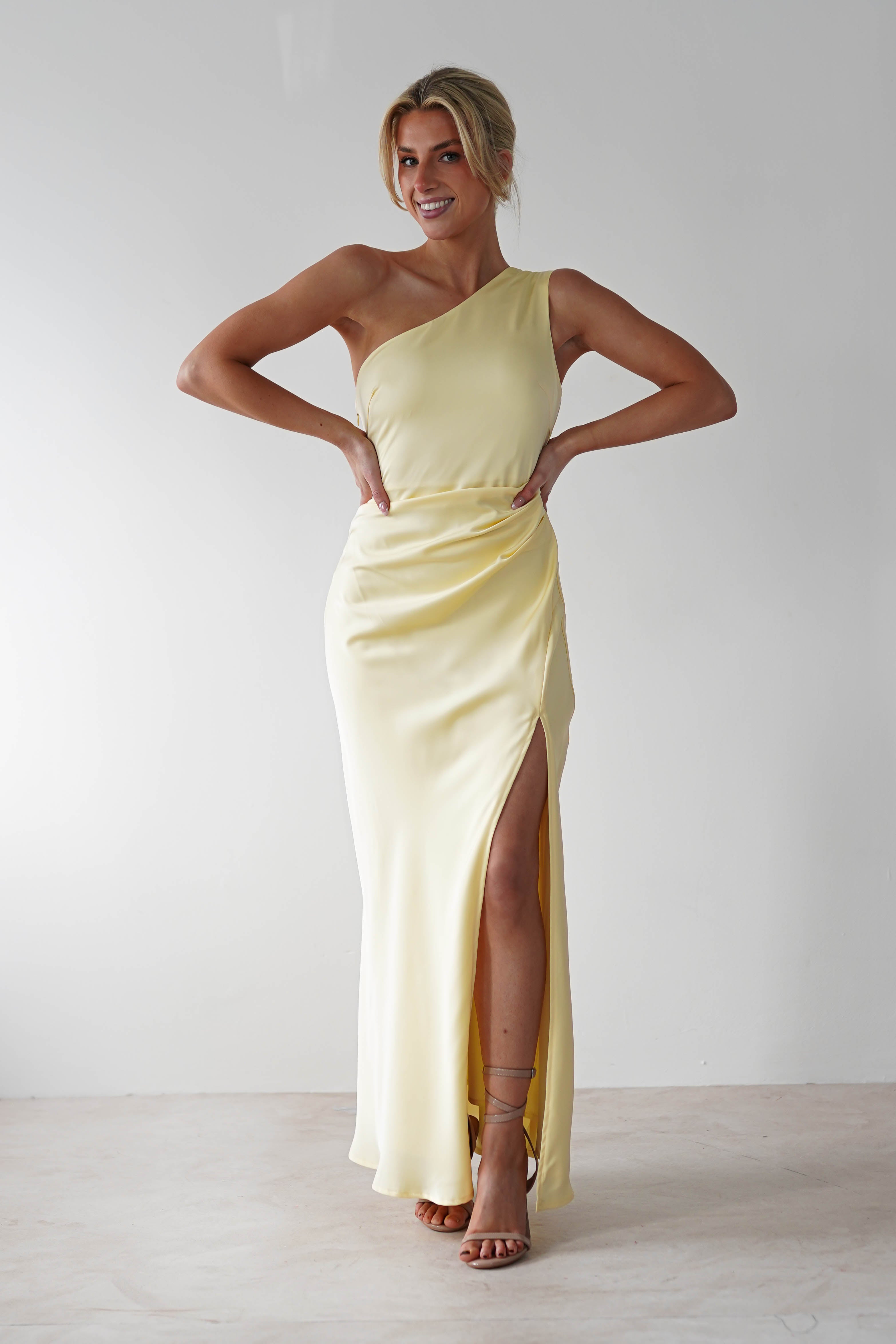 A person in a stunning one-shoulder satin maxi dress with a thigh-high slit stands posing confidently against a plain white background. Their hands are on their hips, and they are wearing brown strappy heels, smiling broadly.