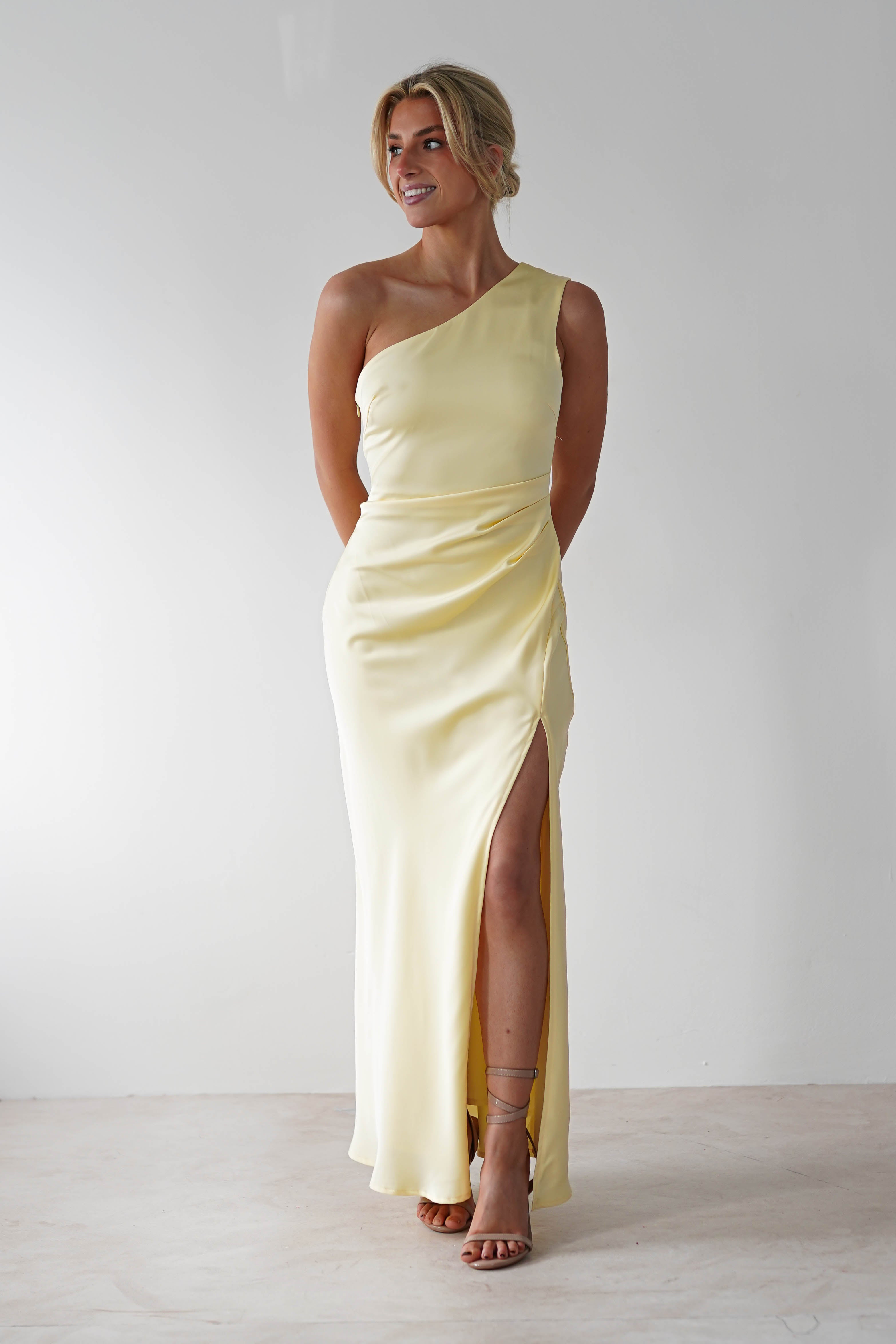 A woman in a pastel yellow, one-shoulder satin maxi dress with a thigh-high slit stands against a plain white background. She is wearing sandals and has her hair styled up, perfect as a wedding guest dress.