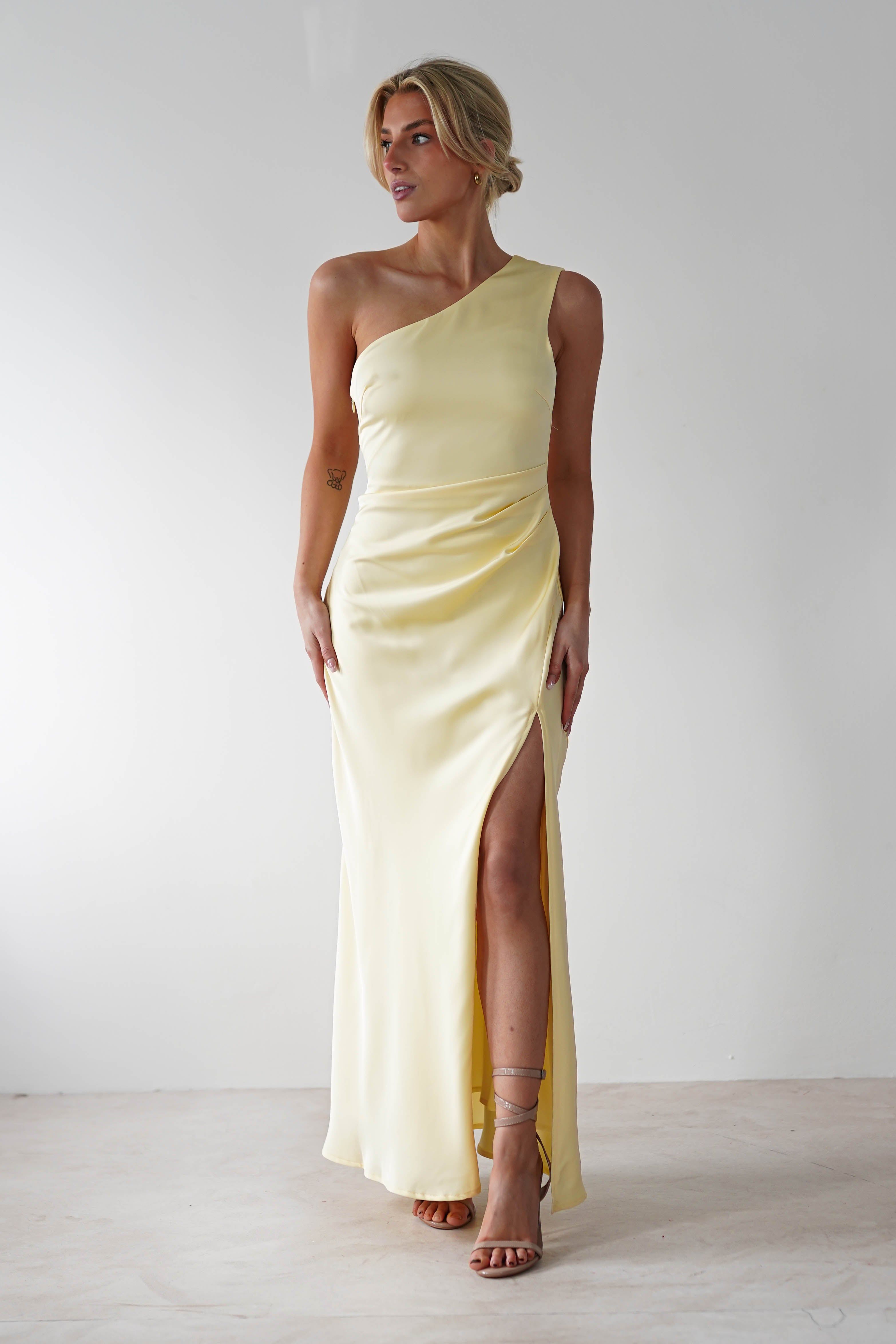 A person stands gracefully in a light yellow, one-shoulder satin maxi dress with a high slit on the left side. They wear strappy heels and have short, styled hair. The plain white background complements their elegant bridal party look.