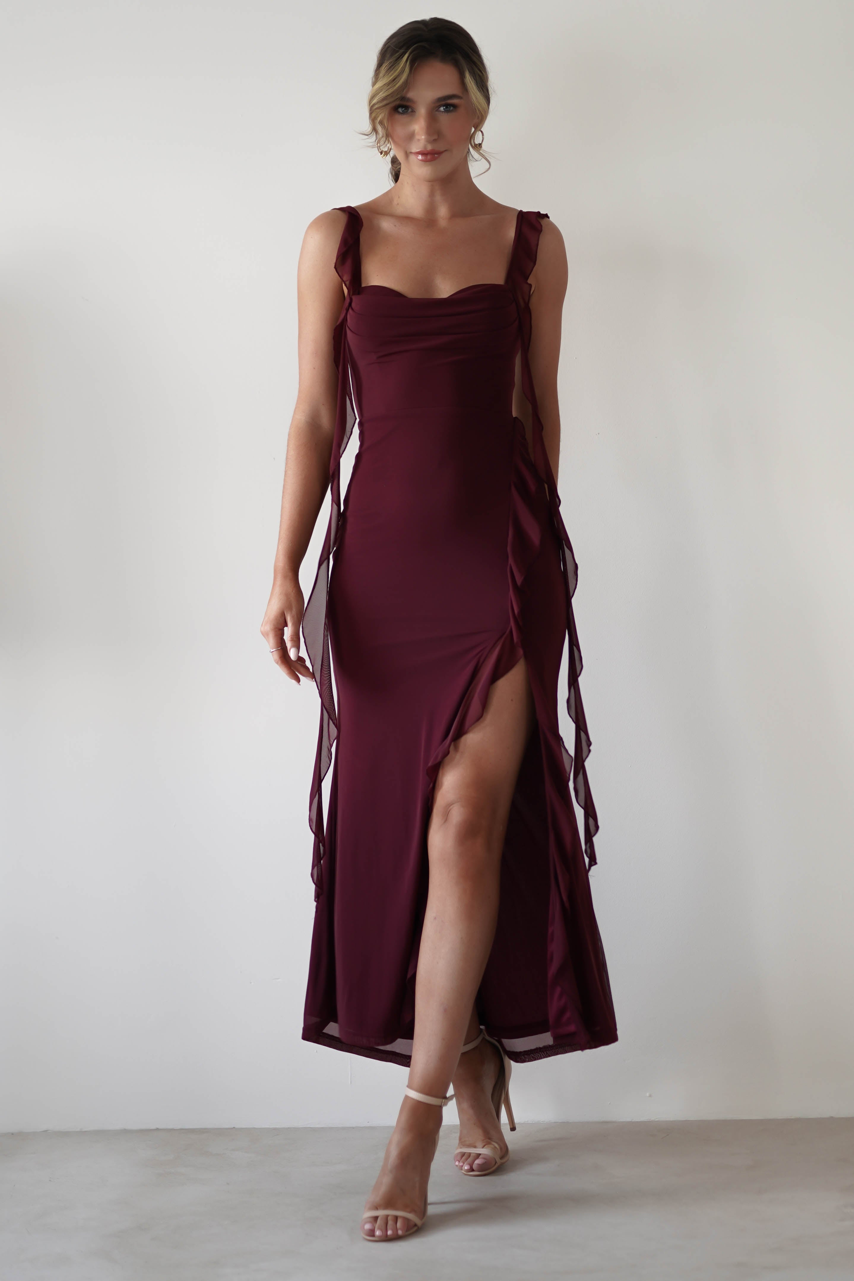 A woman in a sleeveless burgundy dress with chiffon tie straps and a thigh-high slit walks confidently towards the camera. Her high heels complement the elegant look, while her hair styled up completes the ensemble against the plain white backdrop—perfect for a wedding guest.