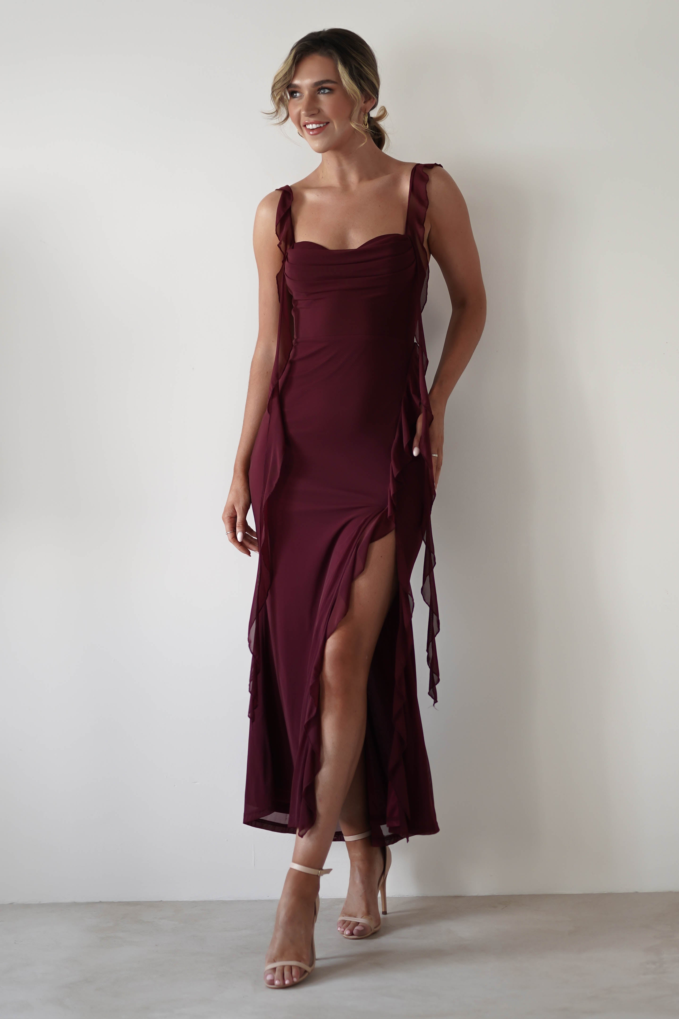 A woman in a burgundy dress with ruffle detail and chiffon tie straps poses against a plain white background. With a stylish thigh-high slit and strappy high heels, shes the perfect picture of elegance—a stunning outfit choice for any wedding guest.