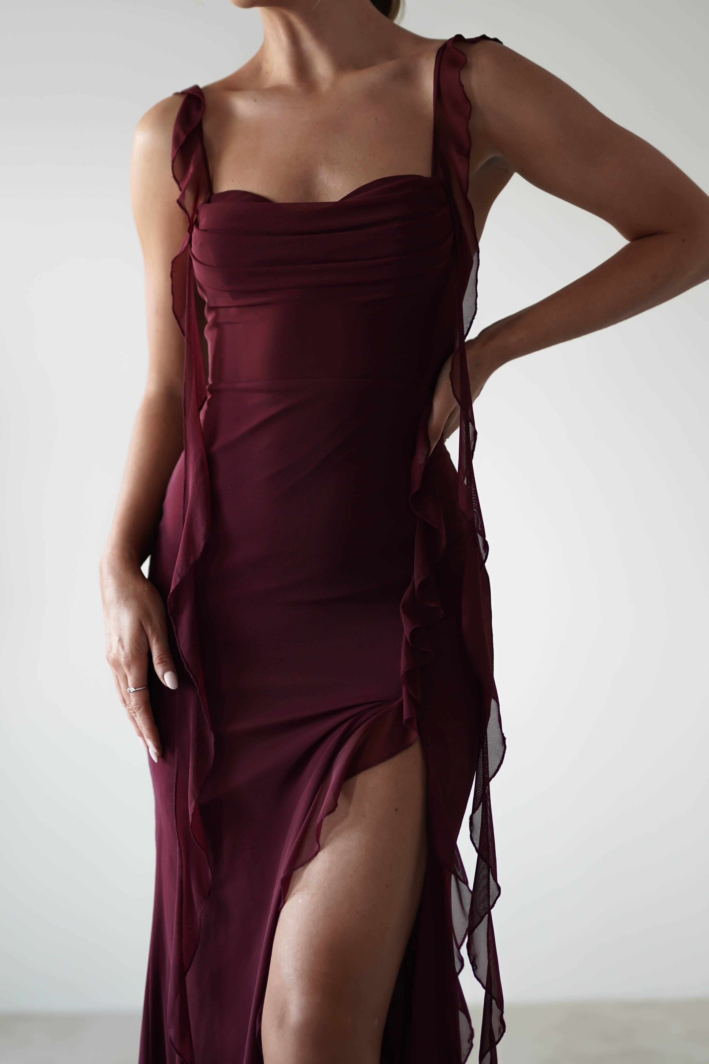 A person in an elegant burgundy dress with chiffon tie straps and a thigh-high slit stands with one hand on their hip. Perfect for a wedding guest, the neutral background enhances the gown’s ruffle details and rich color.