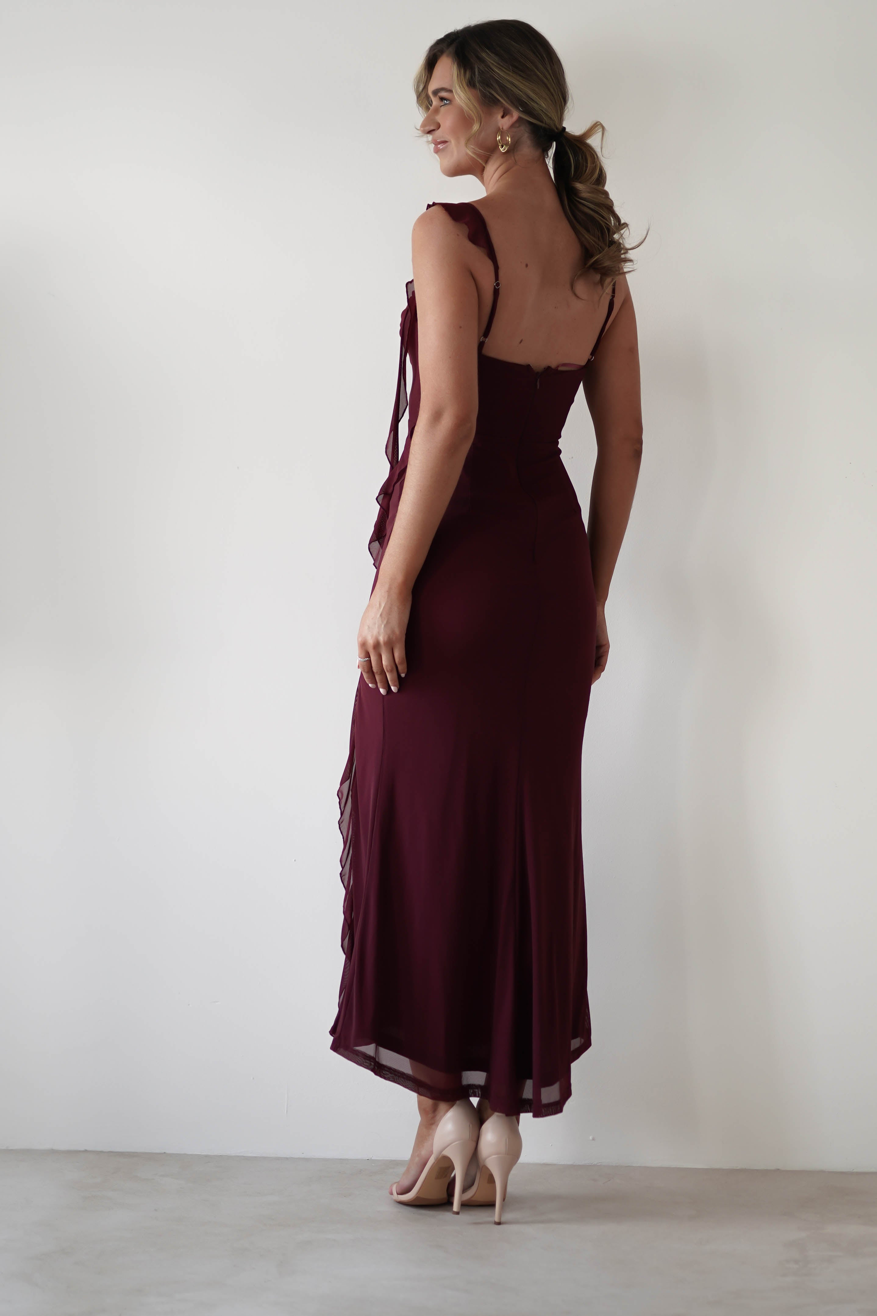 A woman in a sleeveless, dark red dress with chiffon tie straps and ruffle detail stands against a plain light background. Turned slightly away, she showcases the dresss elegant length, perfect for a wedding guest, paired with her light-colored high heels.