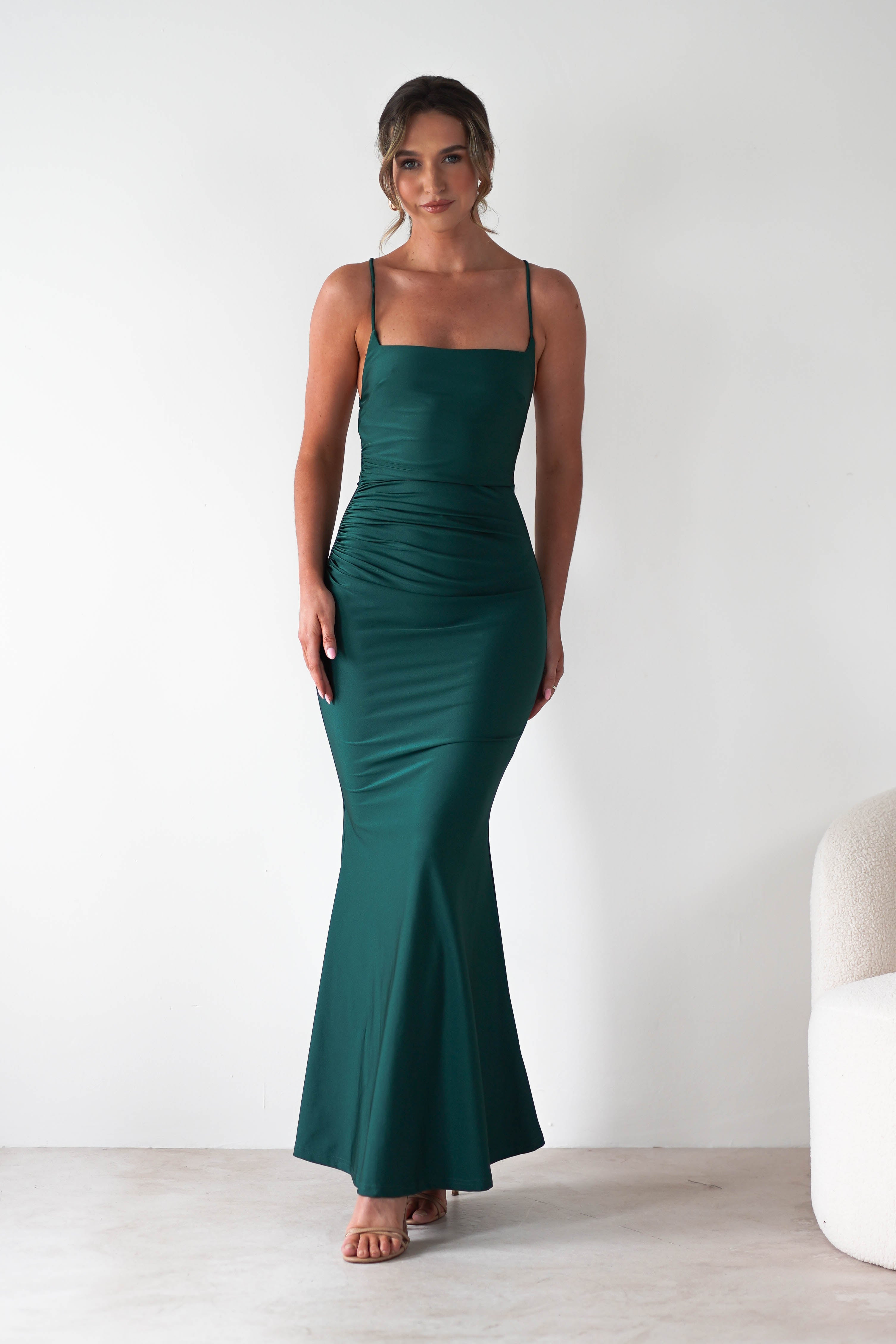 A woman in a fitted, emerald green gown with a mermaid-style skirt and thin straps stands against a plain white background. She has her hair in a loose updo and is wearing nude heels—an elegant look for formal occasions.
