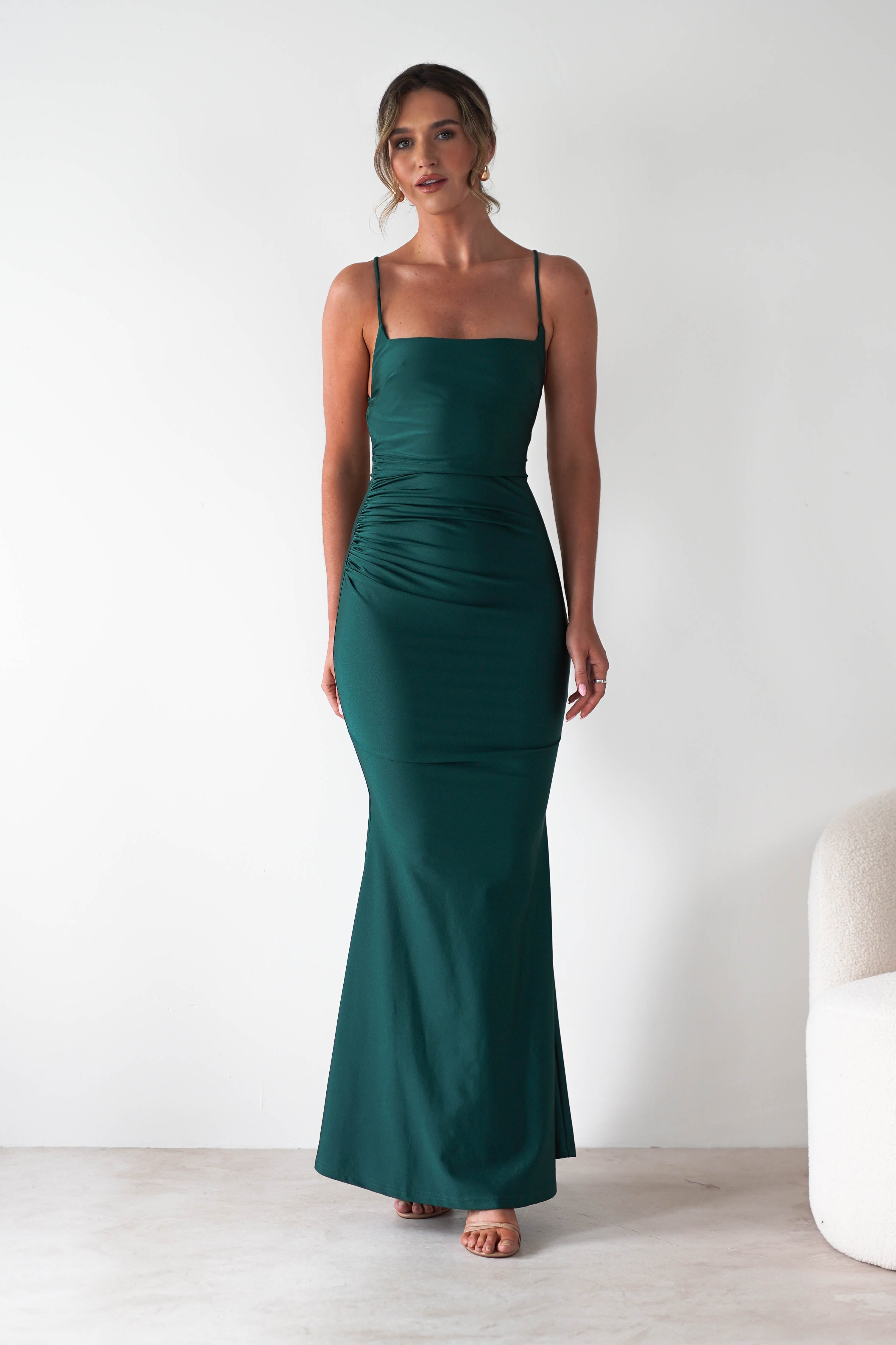 A woman stands indoors in a fitted, floor-length green gown with thin straps and ruched detailing on one side. Featuring a subtle mermaid-style skirt, its perfect for formal occasions. She poses by a white chair against a plain white wall, hair styled in an updo.