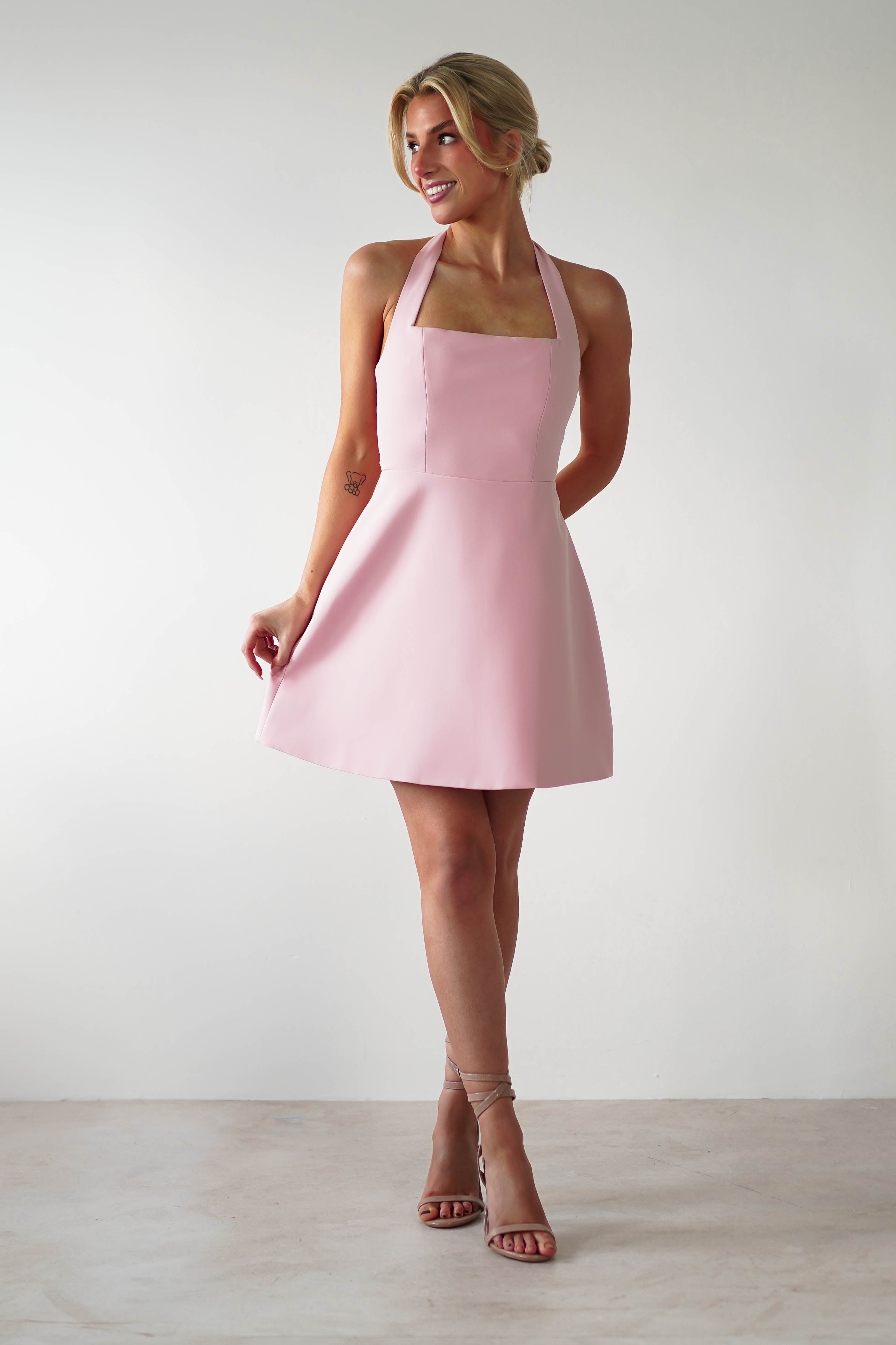 A woman in a pink halterneck mini dress poses elegantly against a neutral background. Slightly angled, she holds the dress gracefully with one hand. Her hair is styled up, and she completes her chic look with strappy nude heels—perfect for wedding guests or parties.