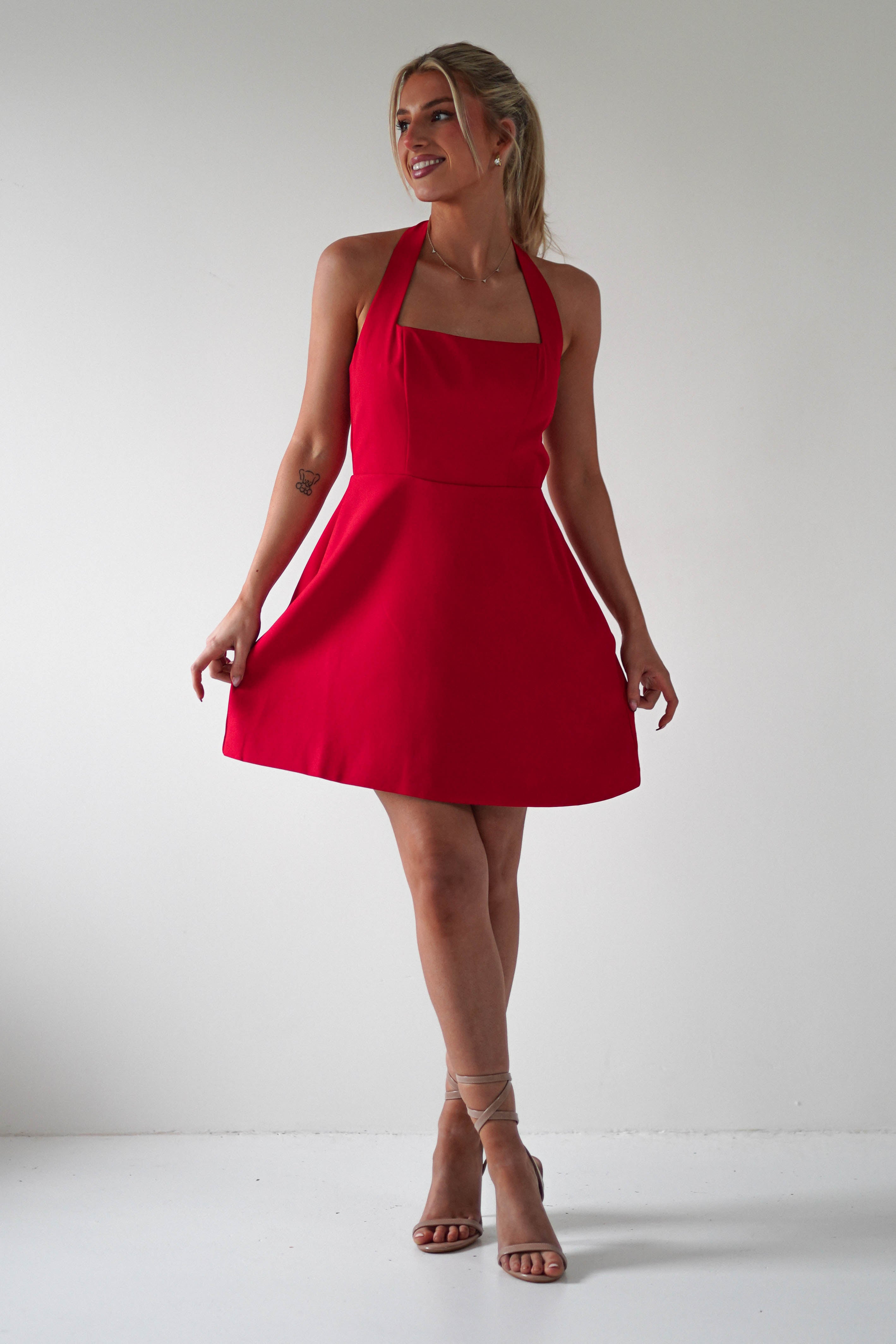 A woman in a chic red halter dress and strappy heels stands against a plain white background, smiling as she holds the sides of her festive Christmas party dress. Her hair is elegantly tied back.