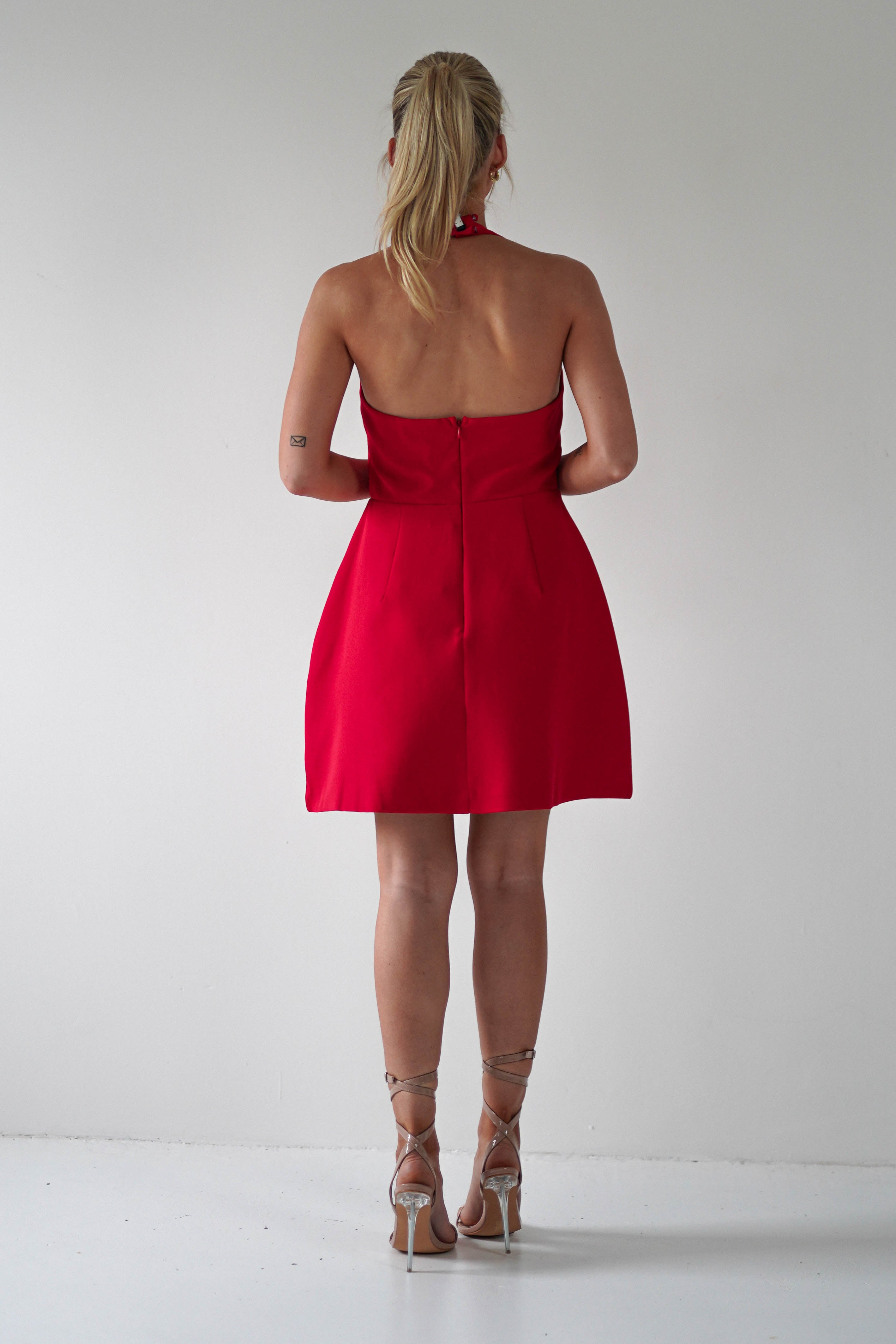 A woman with blonde hair in a ponytail stands facing a white wall. She is wearing a backless, sleeveless red Christmas party dress and strappy high heels. Her arms are by her sides, exuding an air of effortless elegance.