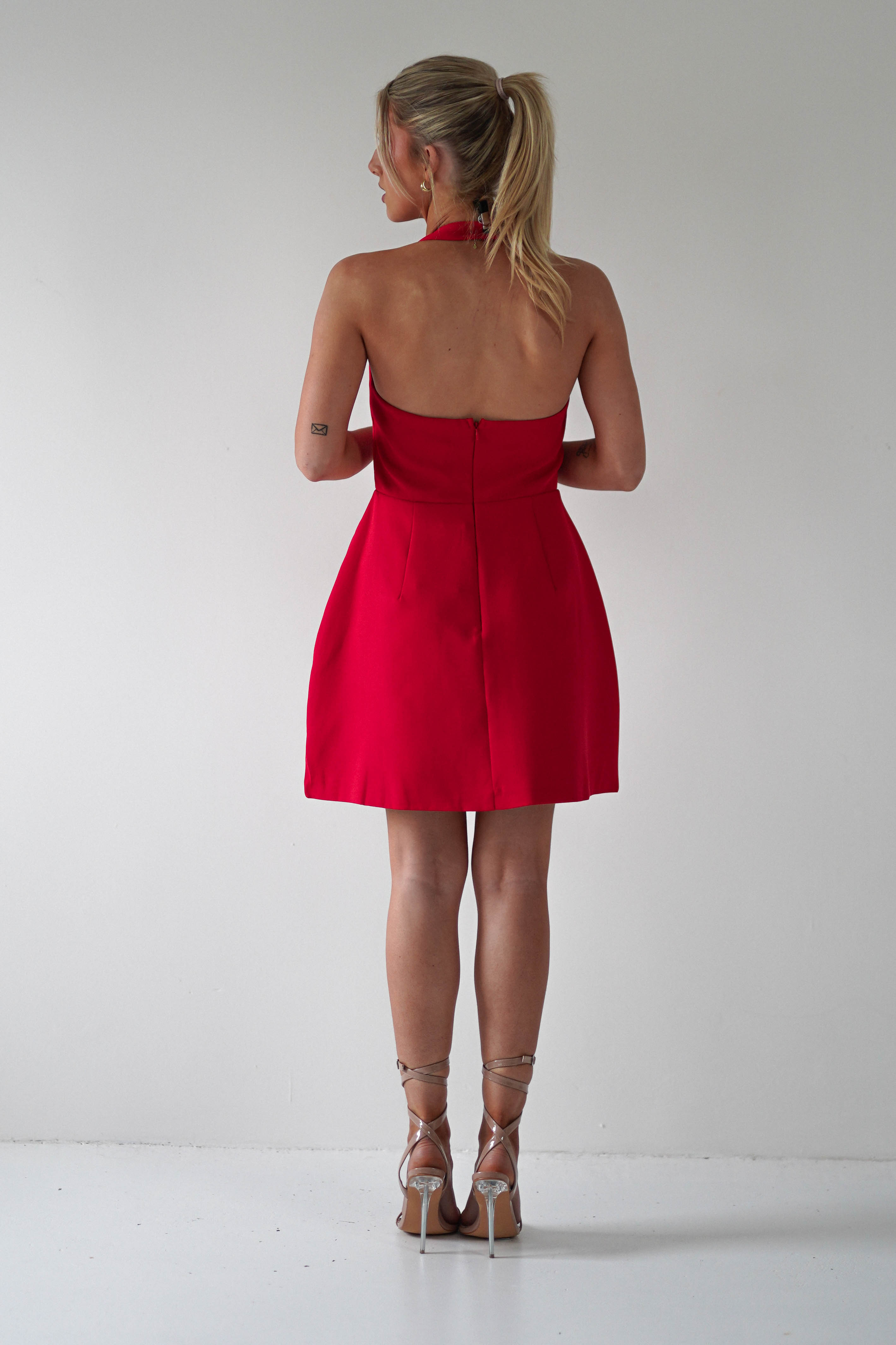 A woman with blonde hair in a ponytail stands facing a white wall, wearing a stunning red Christmas party dress and strappy high heels.