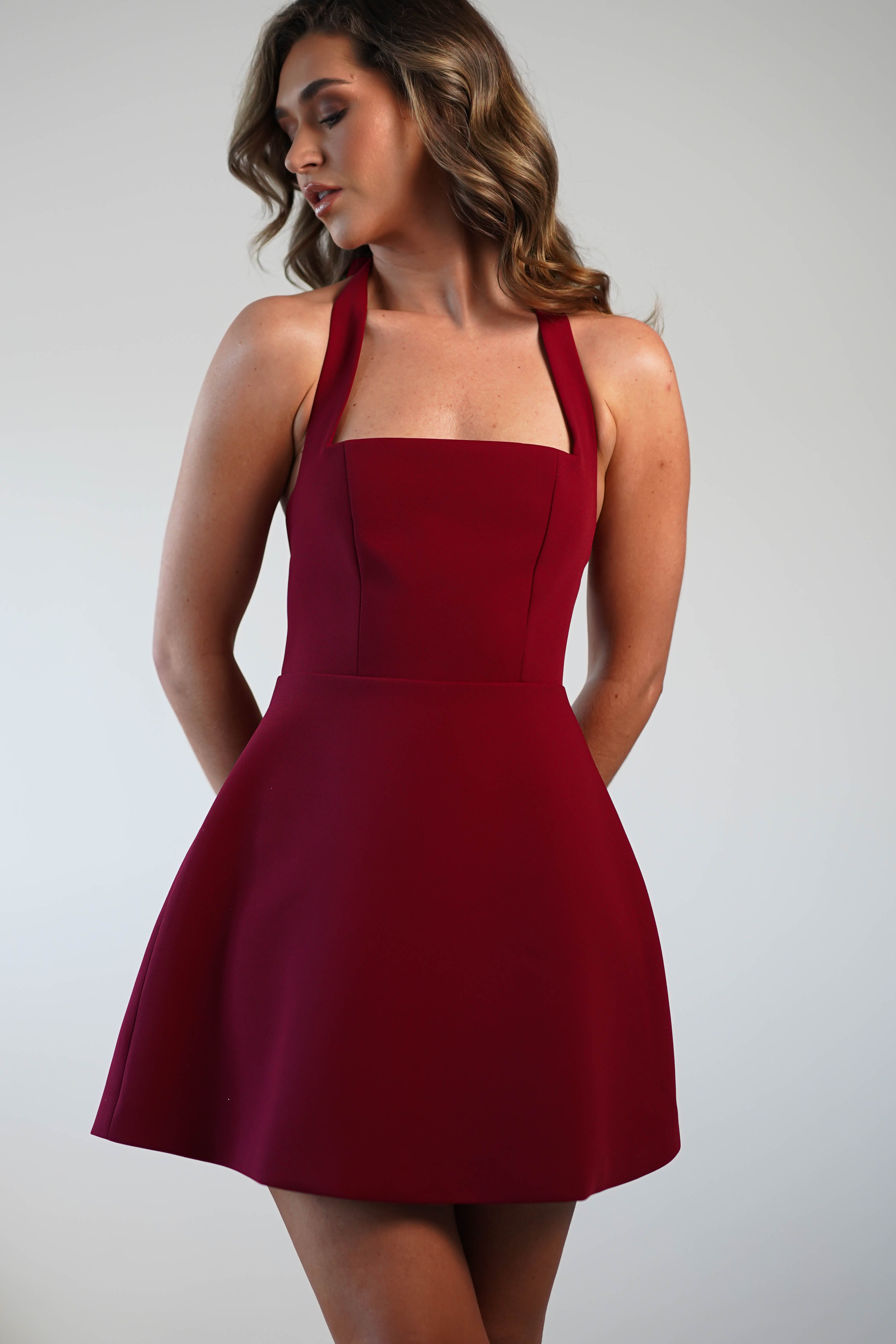 A woman with wavy hair wears a sleeveless, maroon dress with a fitted bodice and flared skirt, perfect for graduations and wedding guest appearances. She looks to the side against a neutral background, her arms relaxed behind her back.