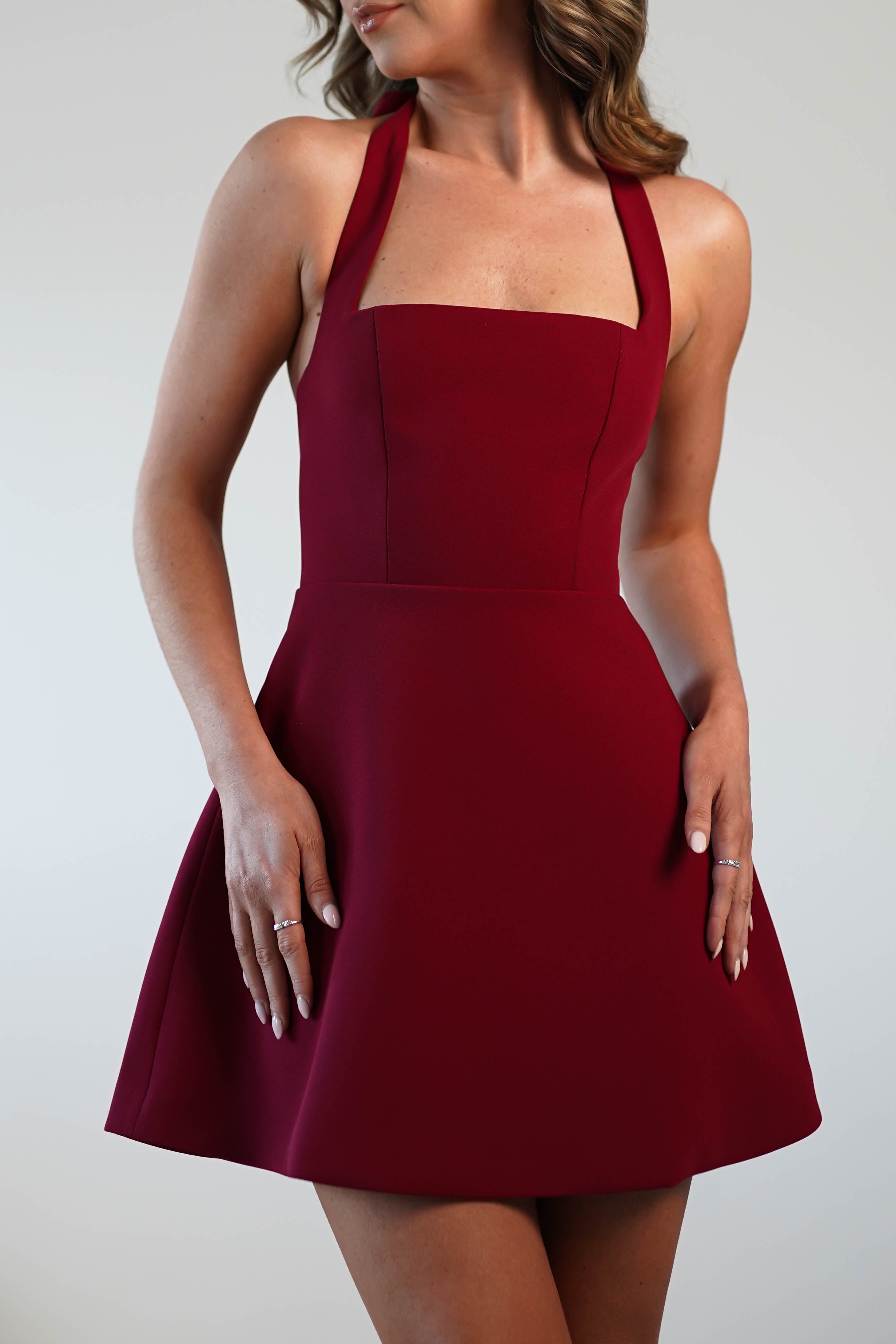 A person elegantly models a sleeveless, deep red dress featuring a square neckline and fitted bodice. Perfect for wedding guests or graduations, the dress boasts a flared skirt against a plain white background.