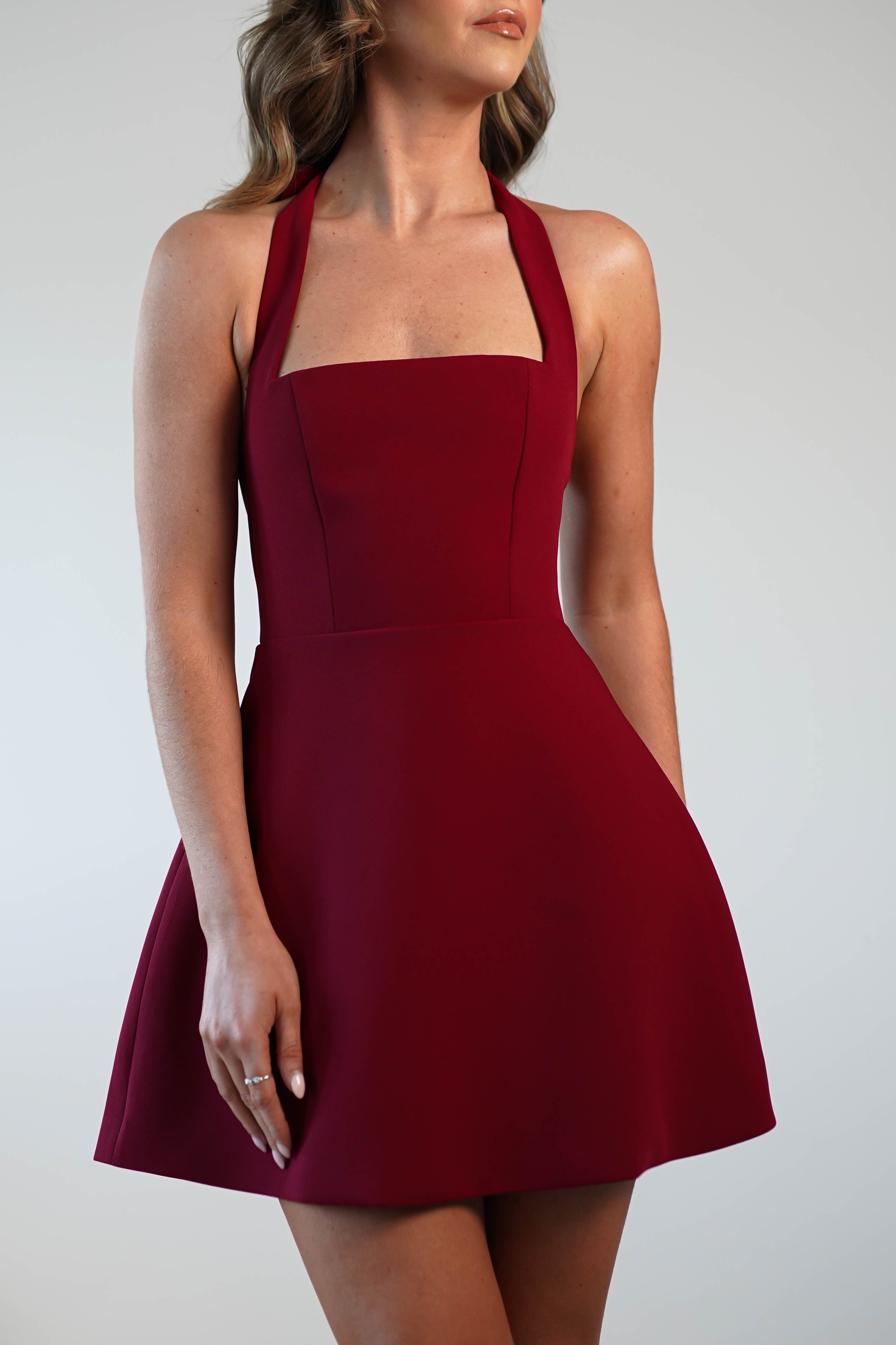 A woman stands against a plain backdrop in a sleeveless burgundy dress with a square neckline and flared skirt, perfect for graduations or as a wedding guest ensemble.