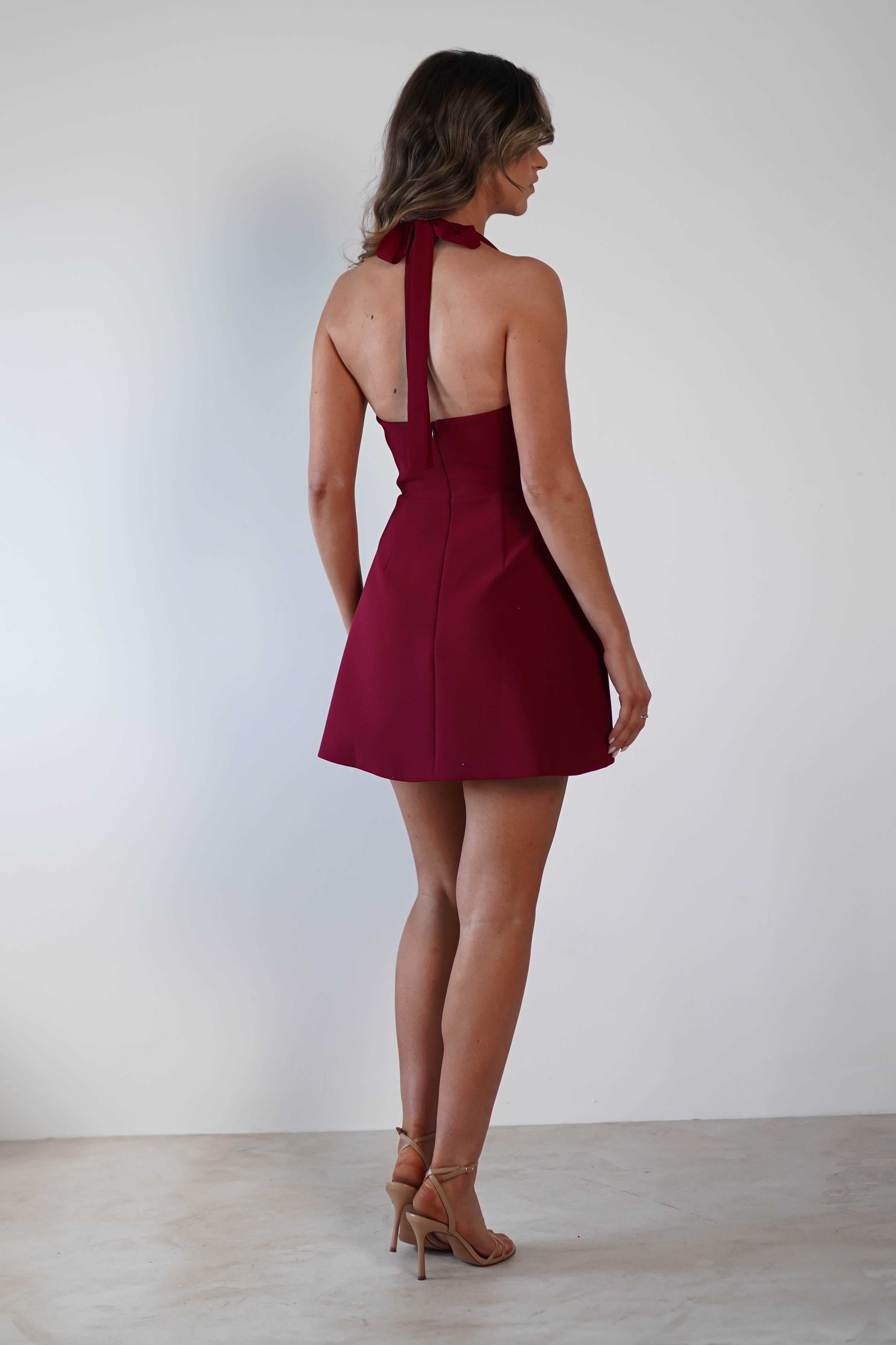 A woman stands with her back to the camera, elegantly poised in a sleeveless, backless halterneck mini burgundy dress. Ideal for a wedding guest or graduations, she completes her look with beige high heels against a plain white background. Her long hair cascades gracefully down her back.