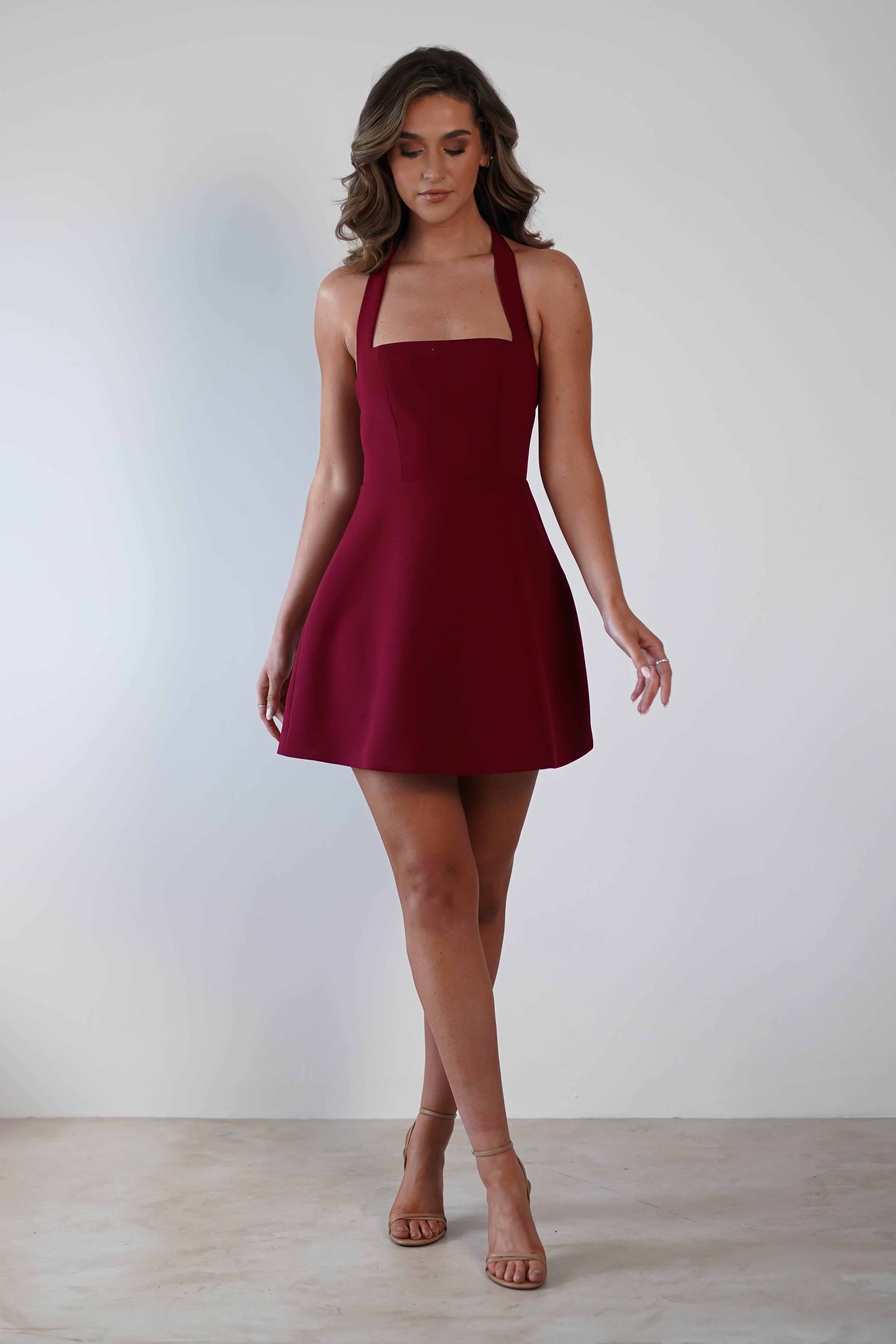 A person in a sleeveless burgundy dress, perfect for a wedding guest, stands confidently in tan high heels against a plain white background. Their hair cascades in loose waves, adding an elegant touch.