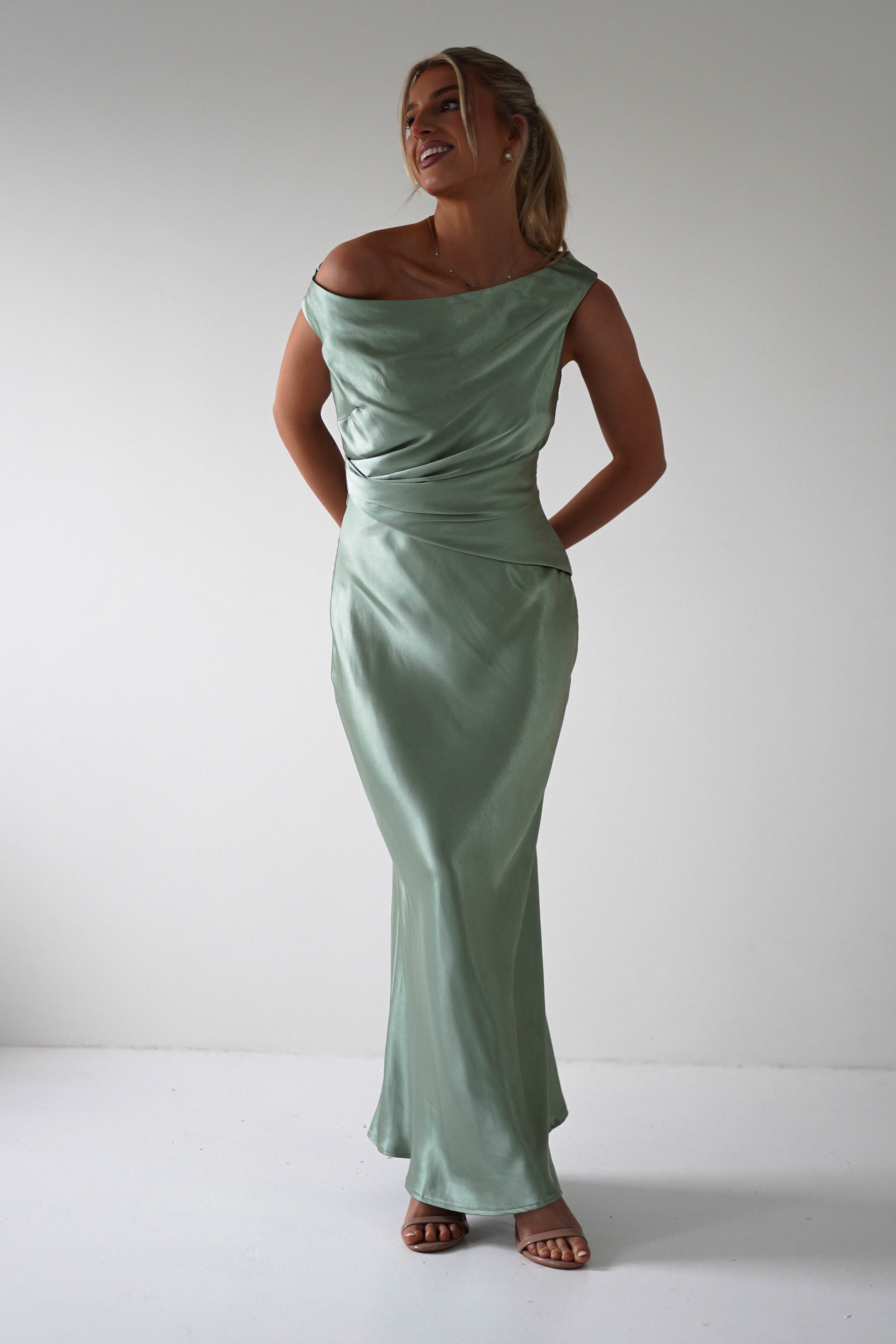 A person is wearing a long, off-shoulder sage green satin dress, perfect for a bridesmaid or wedding guest. Standing against a plain white background, they have their hands behind their back and are smiling. Their hair is tied back, and they complete the look with chic sandals.