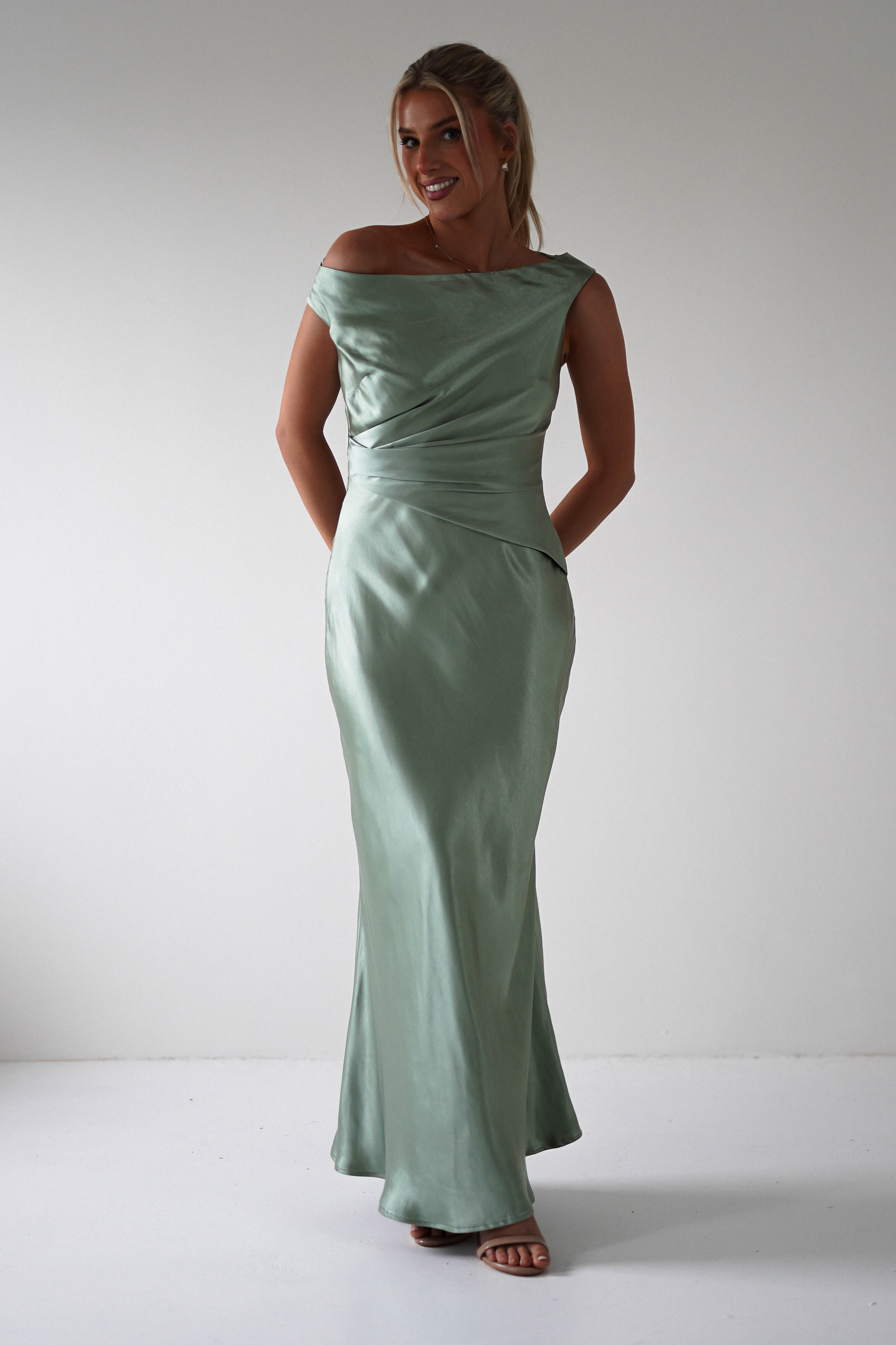 Standing against a plain backdrop, the person wears an elegant, off-the-shoulder, light green satin gown perfect for a wedding guest. The fitted bodice flows gently to the floor, complemented by subtle sandals, evoking a style thats both classic and refined. Their hands rest gracefully behind their back.