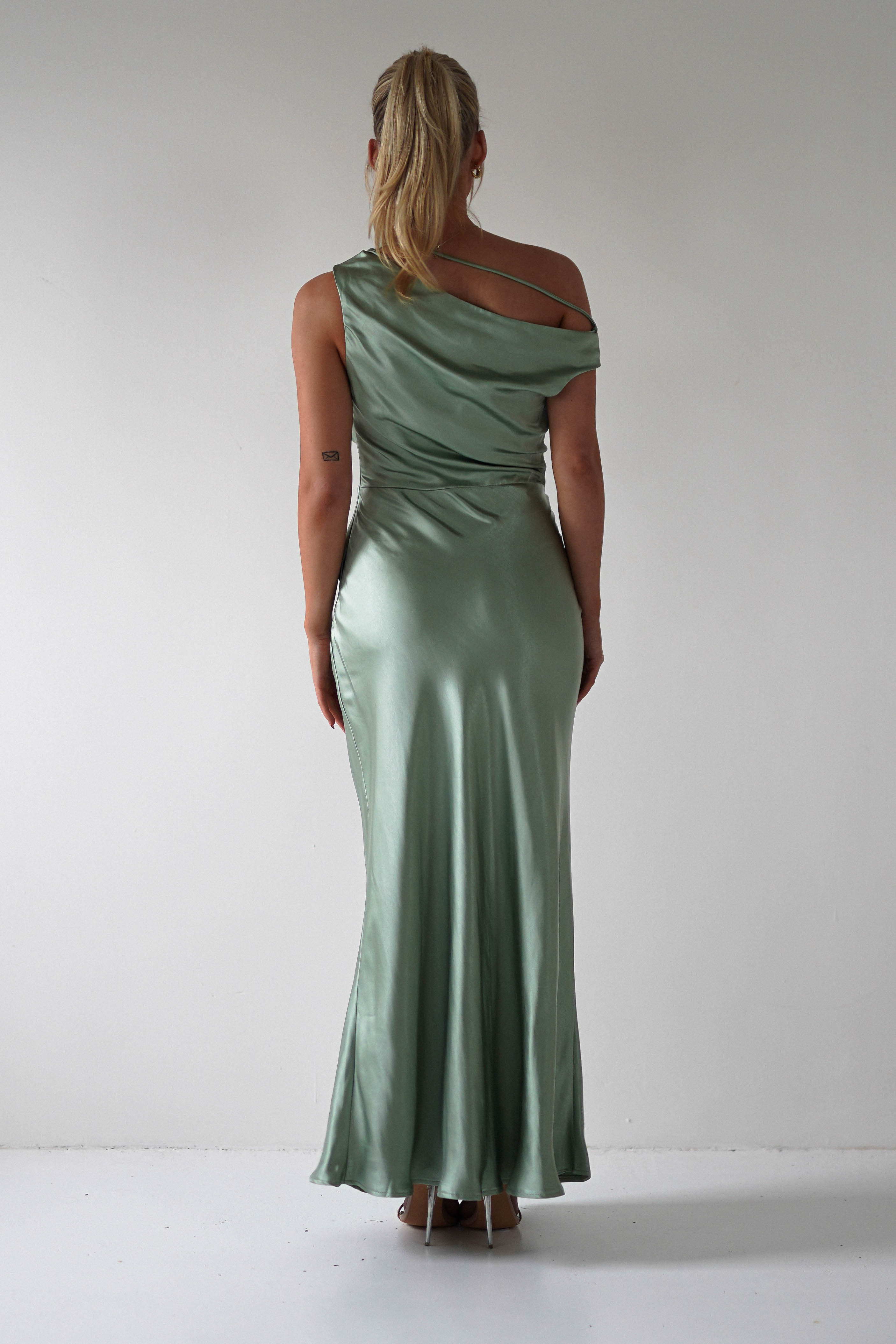 A person with long blonde hair is facing away, wearing a shiny green, one-shoulder gown perfect for a wedding guest. They stand against a plain white background, highlighting the gowns elegant drape and smooth texture.