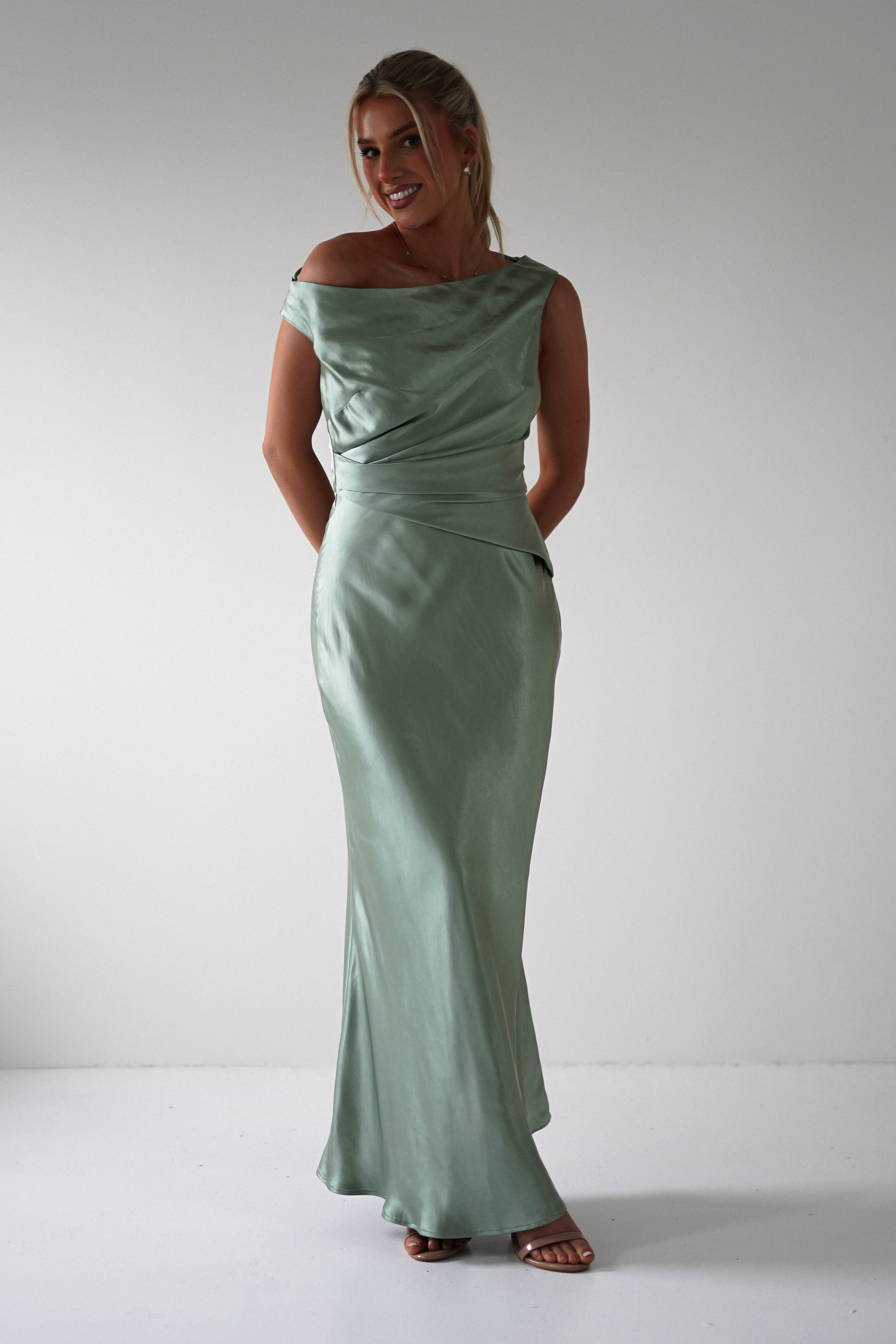 A woman stands in a light green, off-shoulder satin gown with a fitted waist and flowing skirt, perfect for a bridesmaid look. She smiles, looking to the side, with her hands resting in front of her. The background is a plain white wall.