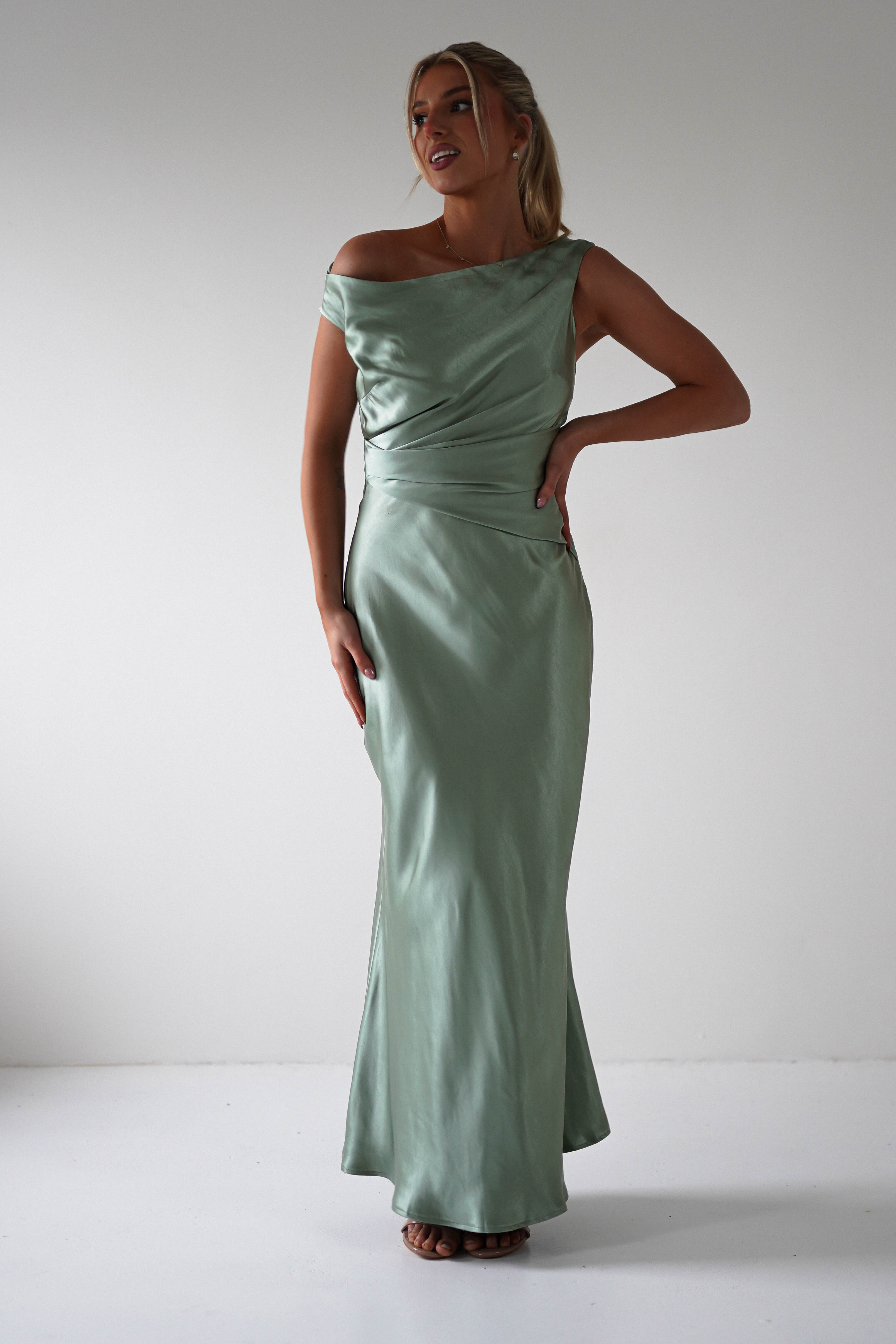 A woman, perhaps a wedding guest or one of the bridesmaids, stands confidently in an elegant, off-the-shoulder, mint green satin gown. Her updo complements the dresss draped design and floor-length cut against a plain background.