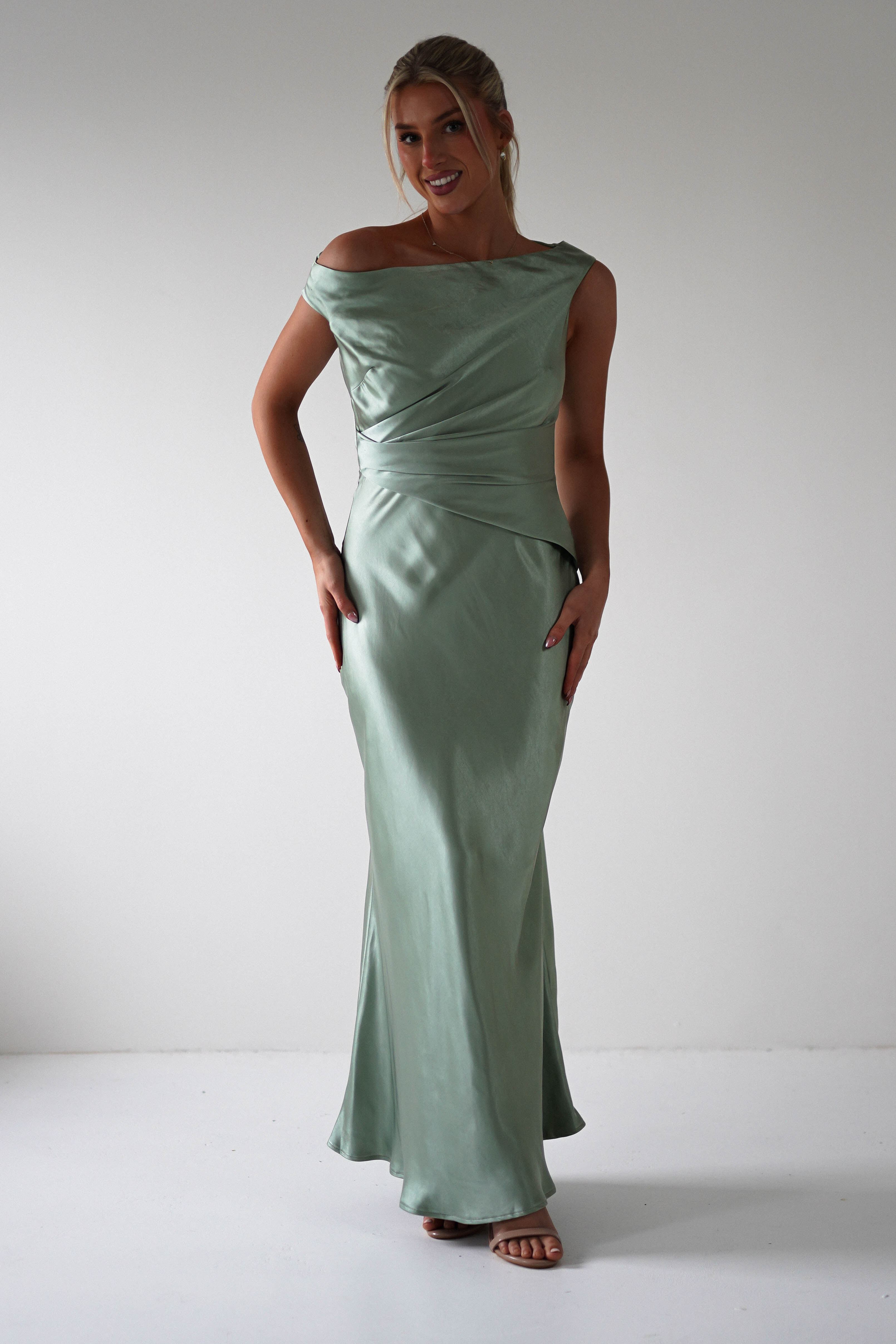 Woman in an elegant, off-shoulder, light green satin gown with a draped design, standing confidently against a plain white background. Perfect for bridesmaids or as a wedding guest, she has her hair styled up, minimal accessories, and is wearing nude sandals.