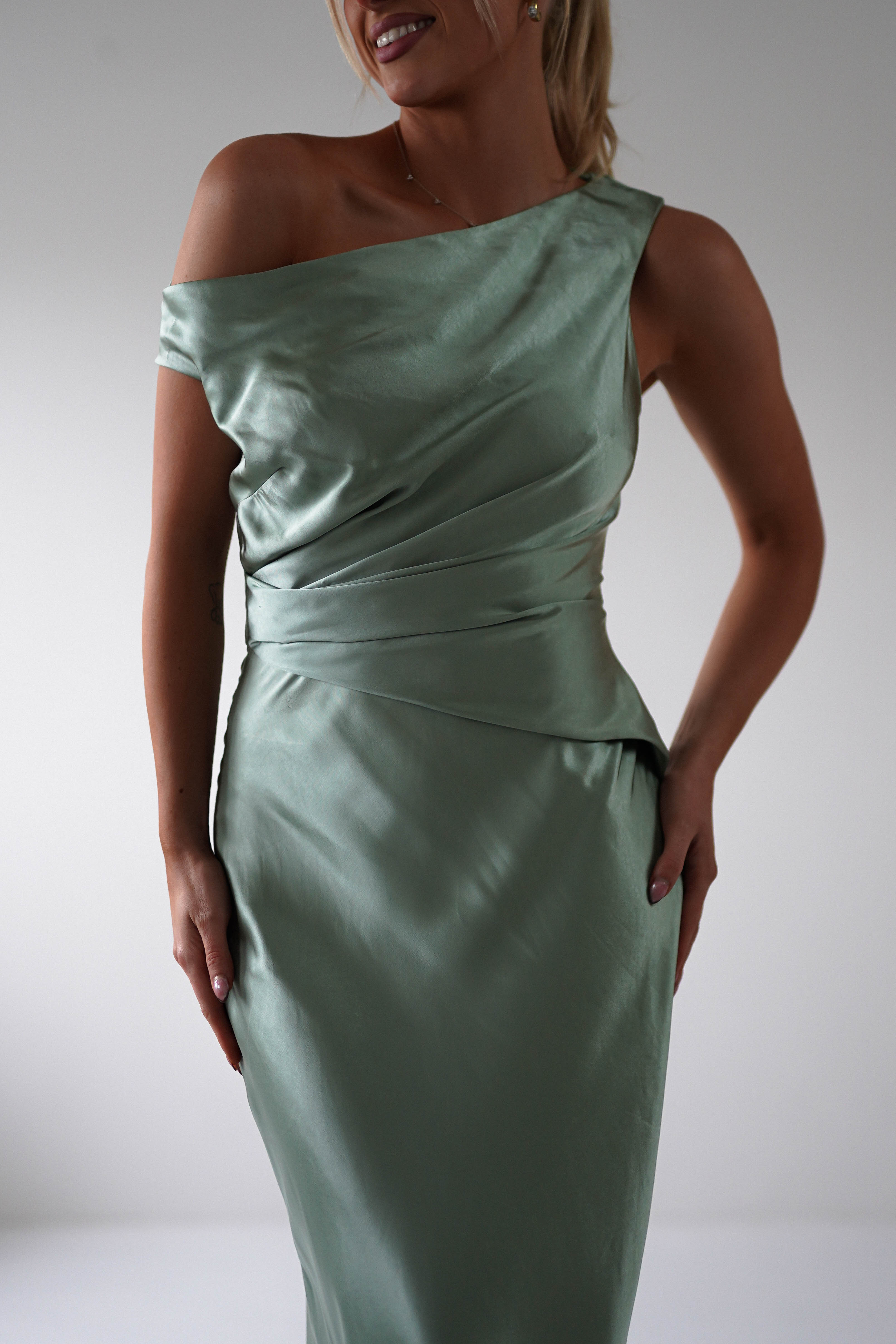 A person wearing a sleek, off-shoulder sage-green satin dress perfect for a wedding guest. The elegant draping and fitted silhouette capture attention, while the neutral background highlights the ensembles charm.