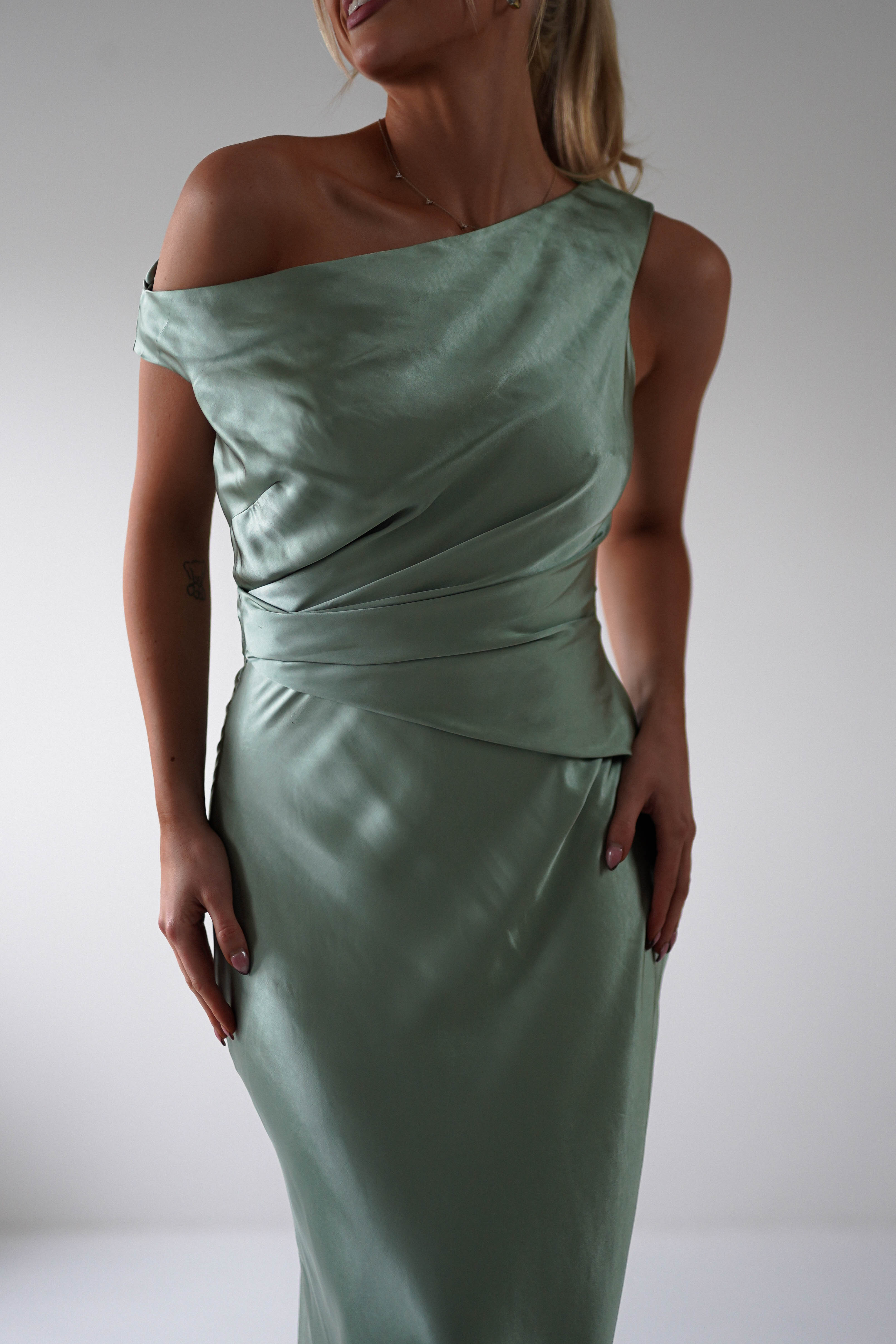 A woman elegantly poses in an off-the-shoulder, light green satin gown, perfect for a wedding guest or bridesmaid, set against a plain backdrop.