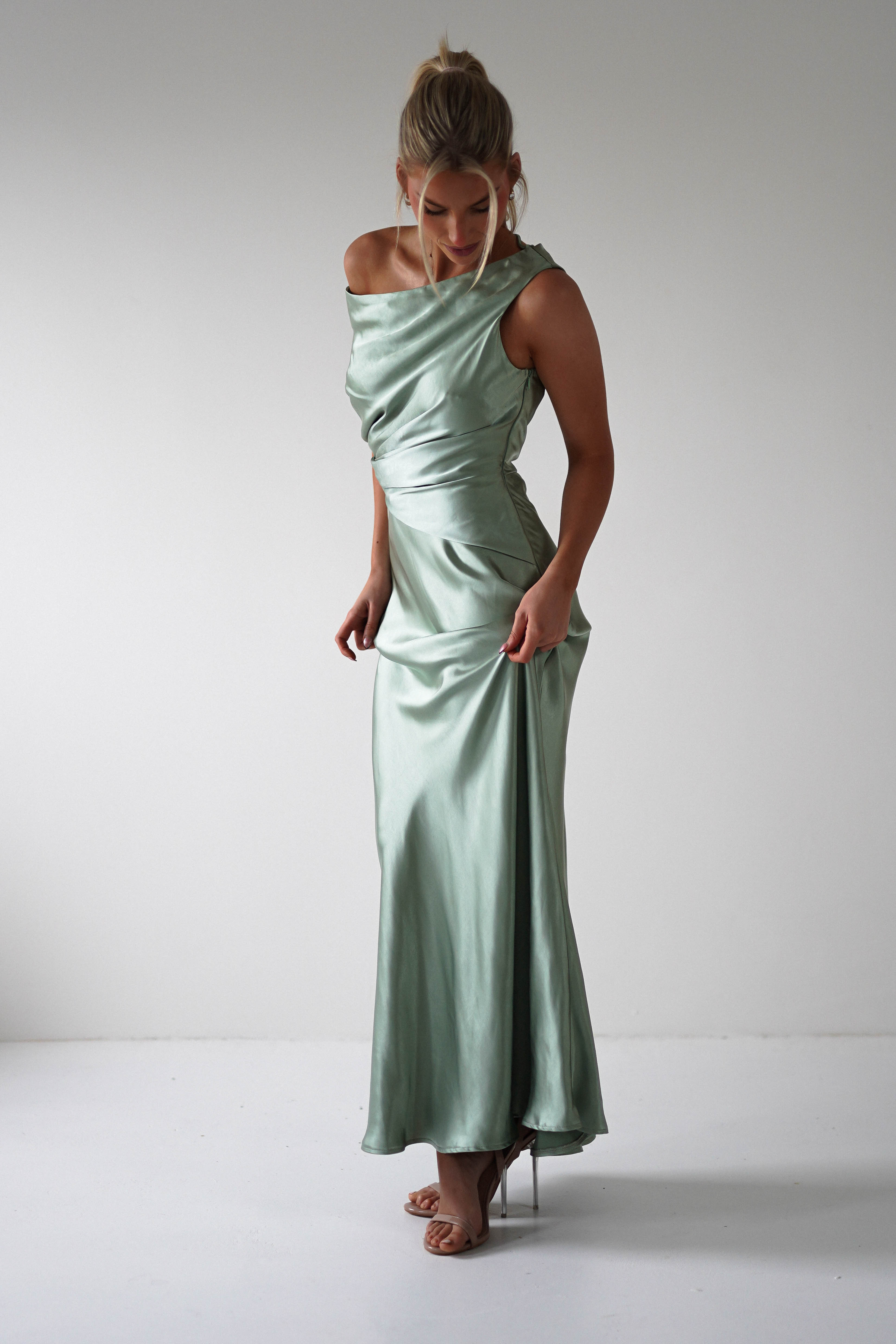 A person wearing a satin, shiny light green gown with an asymmetrical neckline and ruched details stands in a minimalist white room. They are looking down while slightly lifting the dress, perfect attire for a wedding guest.